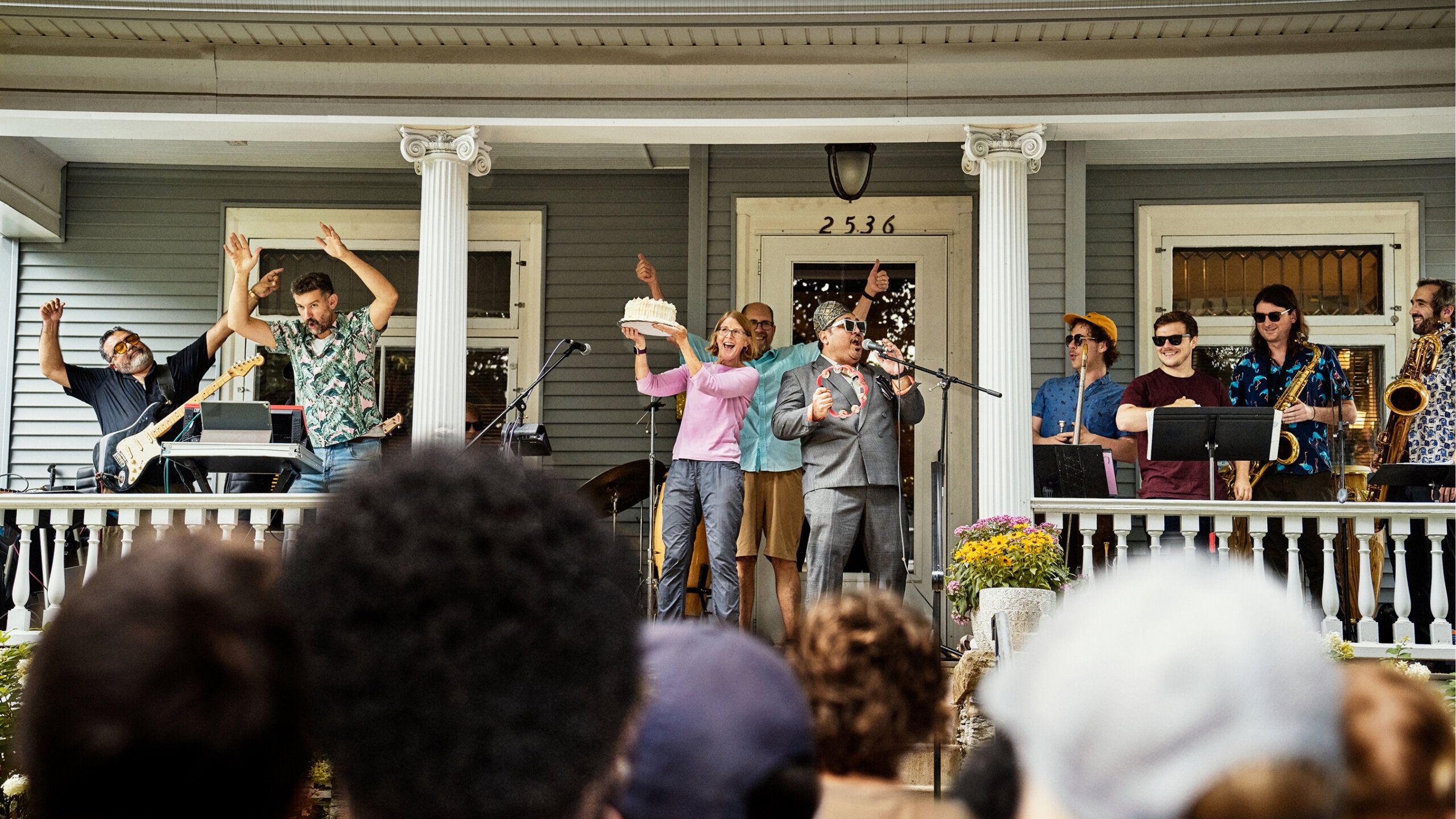 A band performs on a house porch for a crowd. Musicians play instruments and sing, while some people on stage raise their hands. The house number 2536 is visible above the door.
