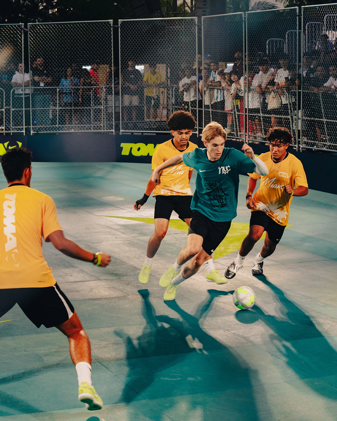 Four young men play soccer on a small, fenced court. Three players in yellow shirts defend against one player in a teal shirt dribbling the ball. Spectators watch from the bleachers behind the fence.