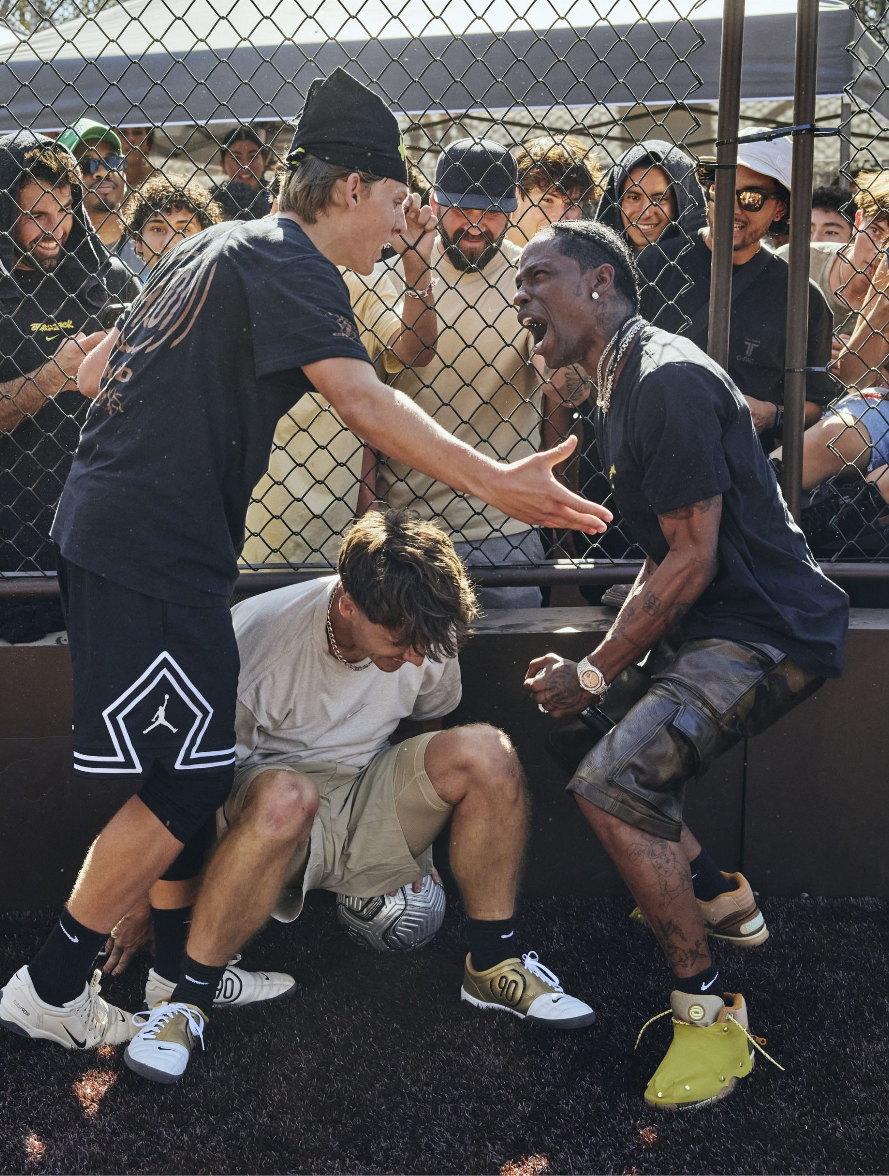 Three men are playing soccer on a fenced court, with two standing and one crouched over the ball; a crowd is watching and reacting in the background.