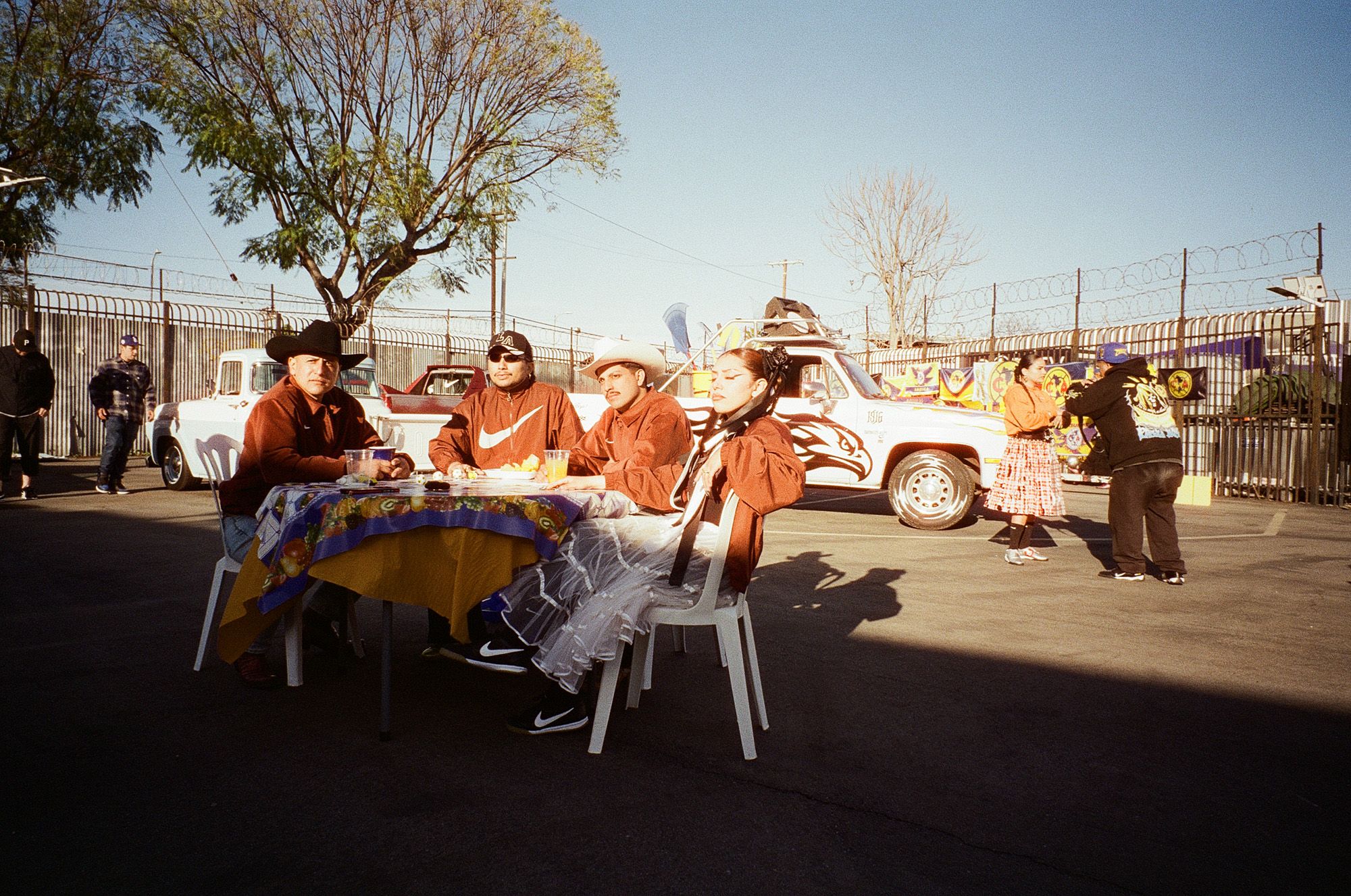 Four people in costumes sit at a table outdoors, with a decorated truck, other people, and trees in the background on a sunny day.
