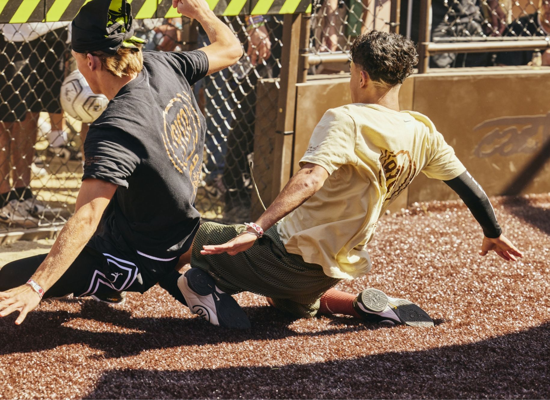 Two young men slide on gravel while competing for a ball during an outdoor sports game, with spectators watching behind a chain-link fence.