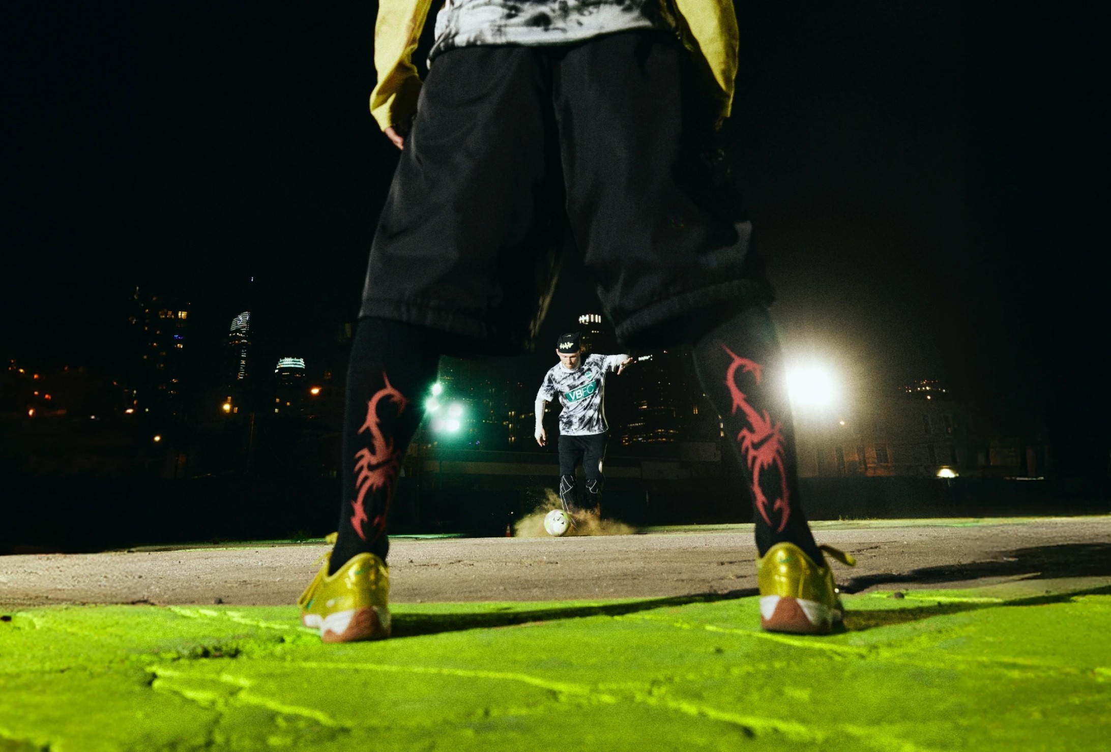 A person stands in the foreground with legs apart while another person kicks a soccer ball in the background at night on a lit urban field.