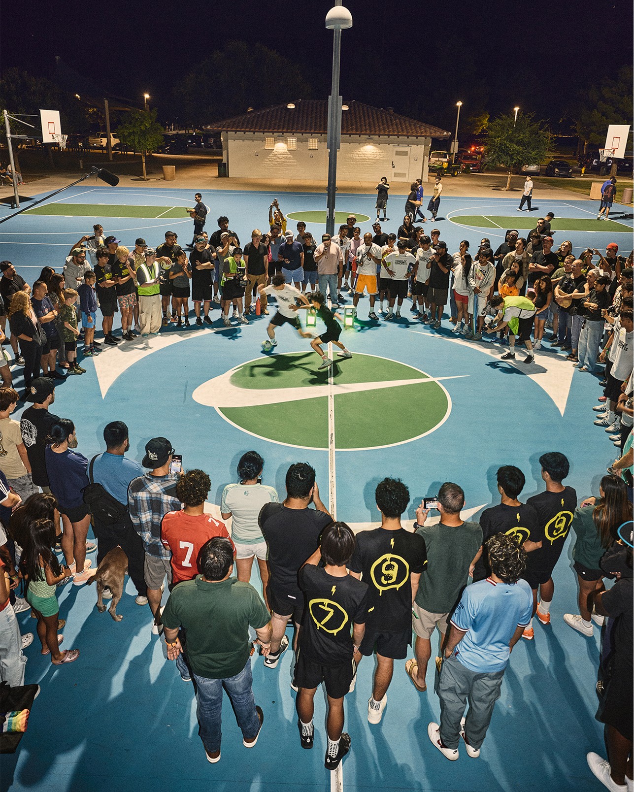 A crowd circles around two people playing basketball on an outdoor court at night, with spectators closely watching the game.