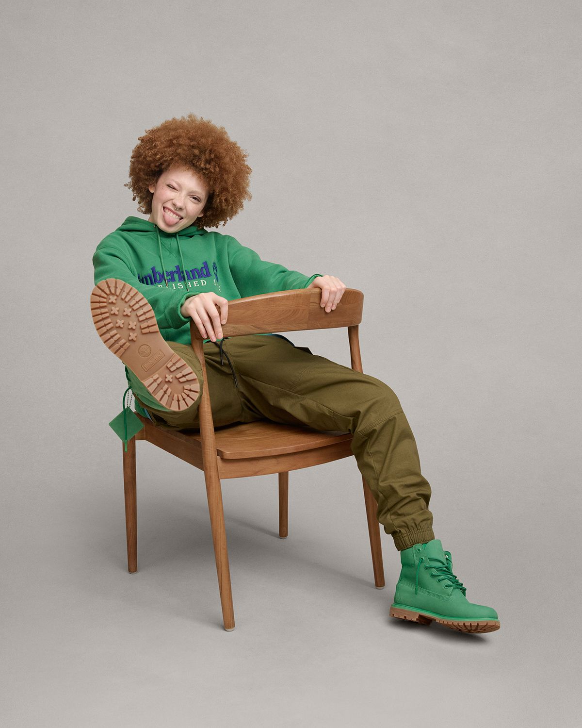 A person sitting on a wooden chair with a green sweatshirt kicking one leg in air. They are wearing green Timberland boots