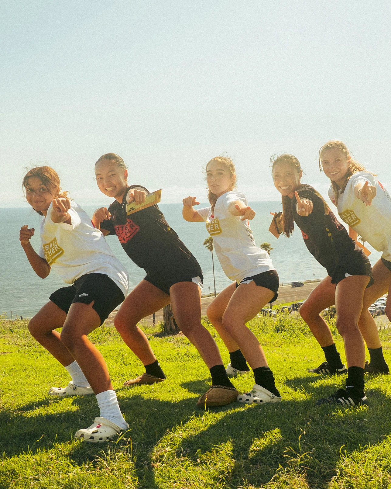 Five girls in athletic wear pose in a crouched position on grass near the ocean, pointing toward the camera with smiles on their faces.