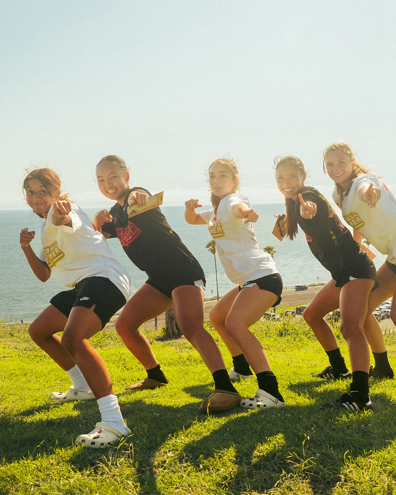 Five girls in athletic wear pose in a crouched position on grass near the ocean, pointing toward the camera with smiles on their faces.