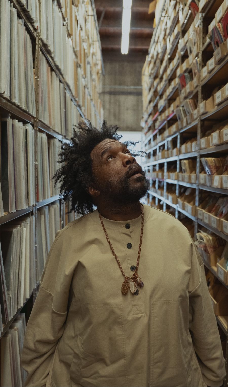 A man with a beard and necklace stands between shelves filled with records, looking upward in a well-lit storage or archive room.
