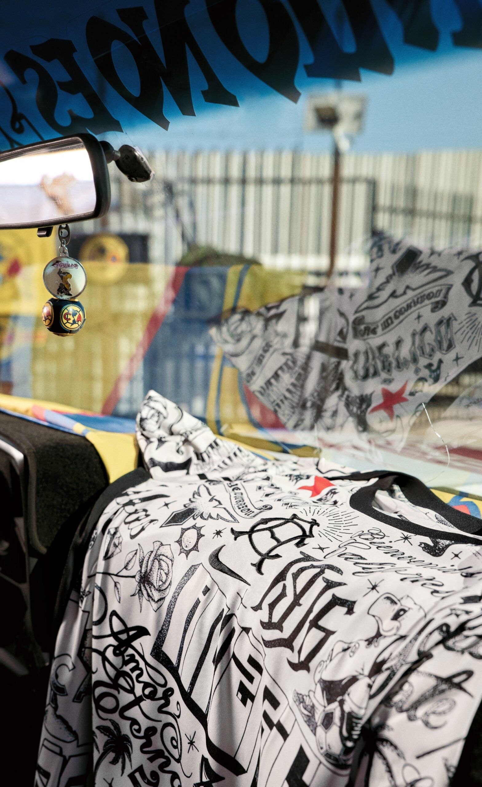 Black and white graphic T-shirt draped over a car dashboard, with cartoon-themed air fresheners hanging from the rearview mirror and a colorful fence visible outside the window.