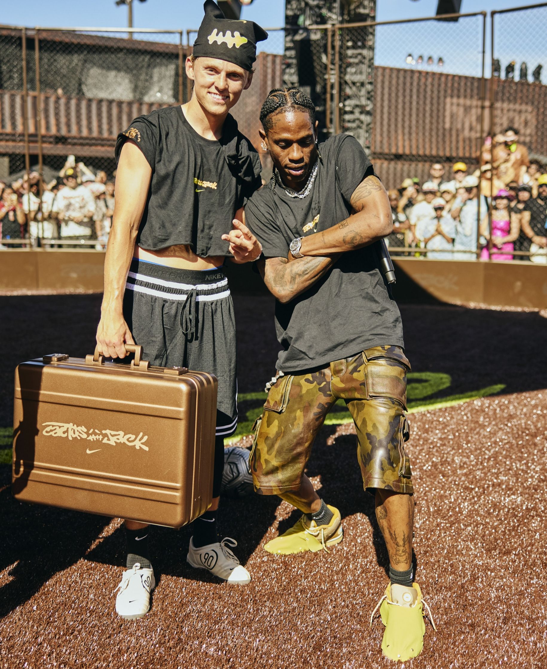 Two men wearing casual streetwear pose together outdoors; one holds a brown Cactus Jack Nike briefcase, and a crowd is visible in the background.