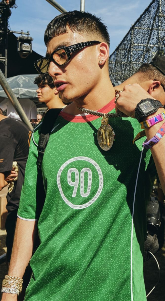 A person wearing a green "90" jersey, large sunglasses, and several bracelets and necklaces stands outdoors at a crowded event.