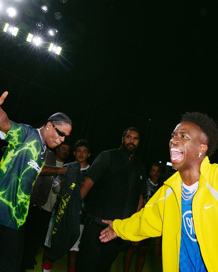 Two men in colorful outfits stand in a crowd under bright lights, one with arms outstretched and another laughing, with people visible in the background.