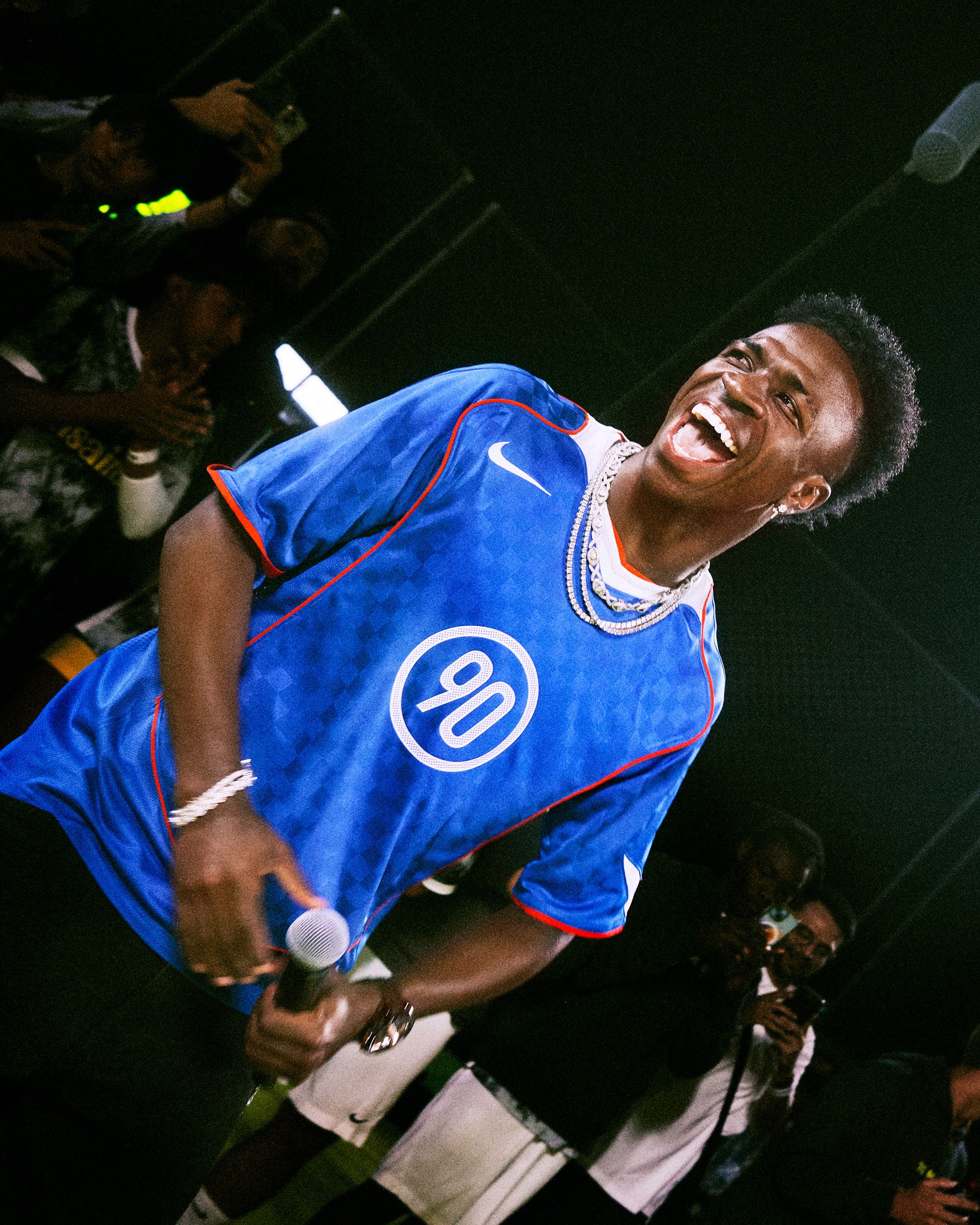 A man wearing a blue sports jersey and holding a microphone smiles broadly while standing in front of an audience.