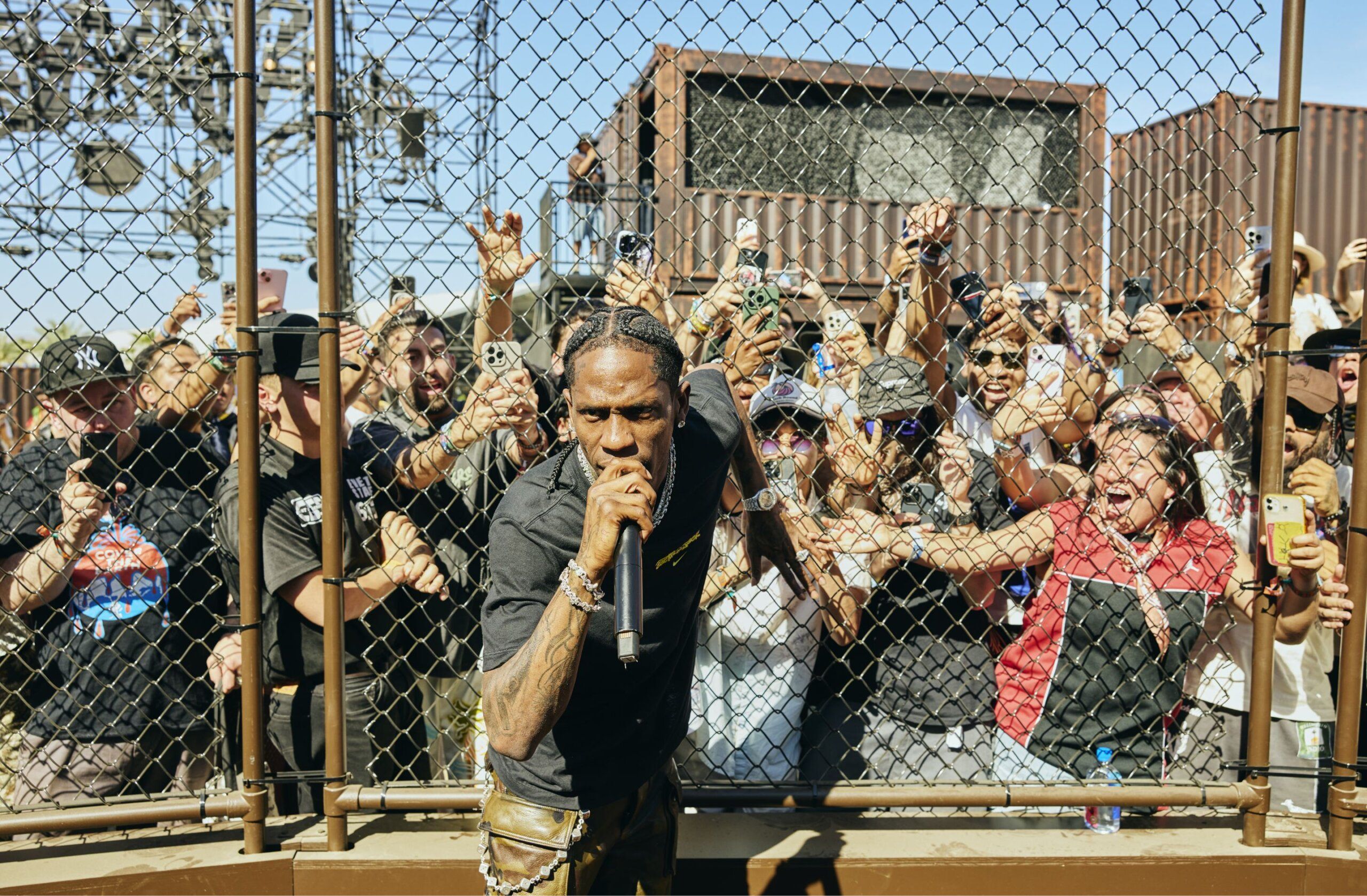 A performer sings into a microphone in front of a chain-link fence, with a large crowd of fans reaching out and taking photos behind the fence.