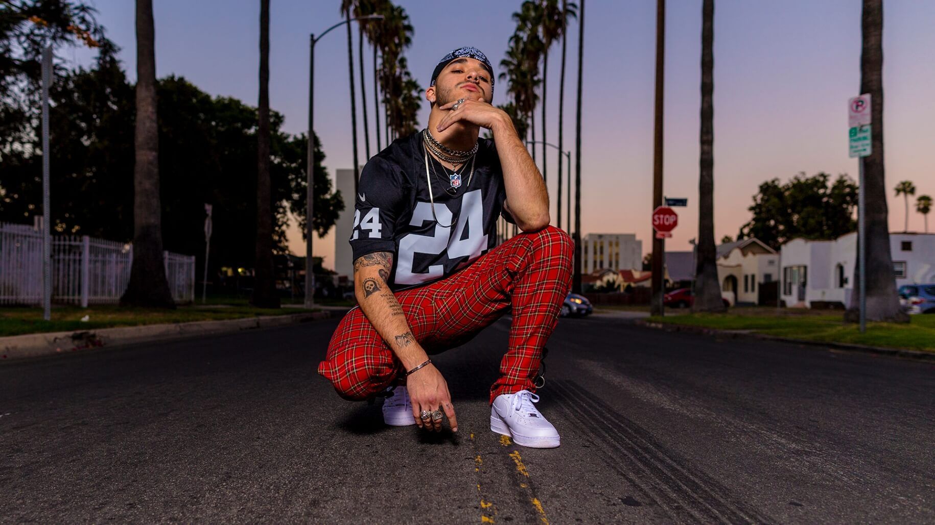Man kneeling posing in palm tree lined street at dusk wearing number 24 Raiders football jersey, necklaces and plaid pants