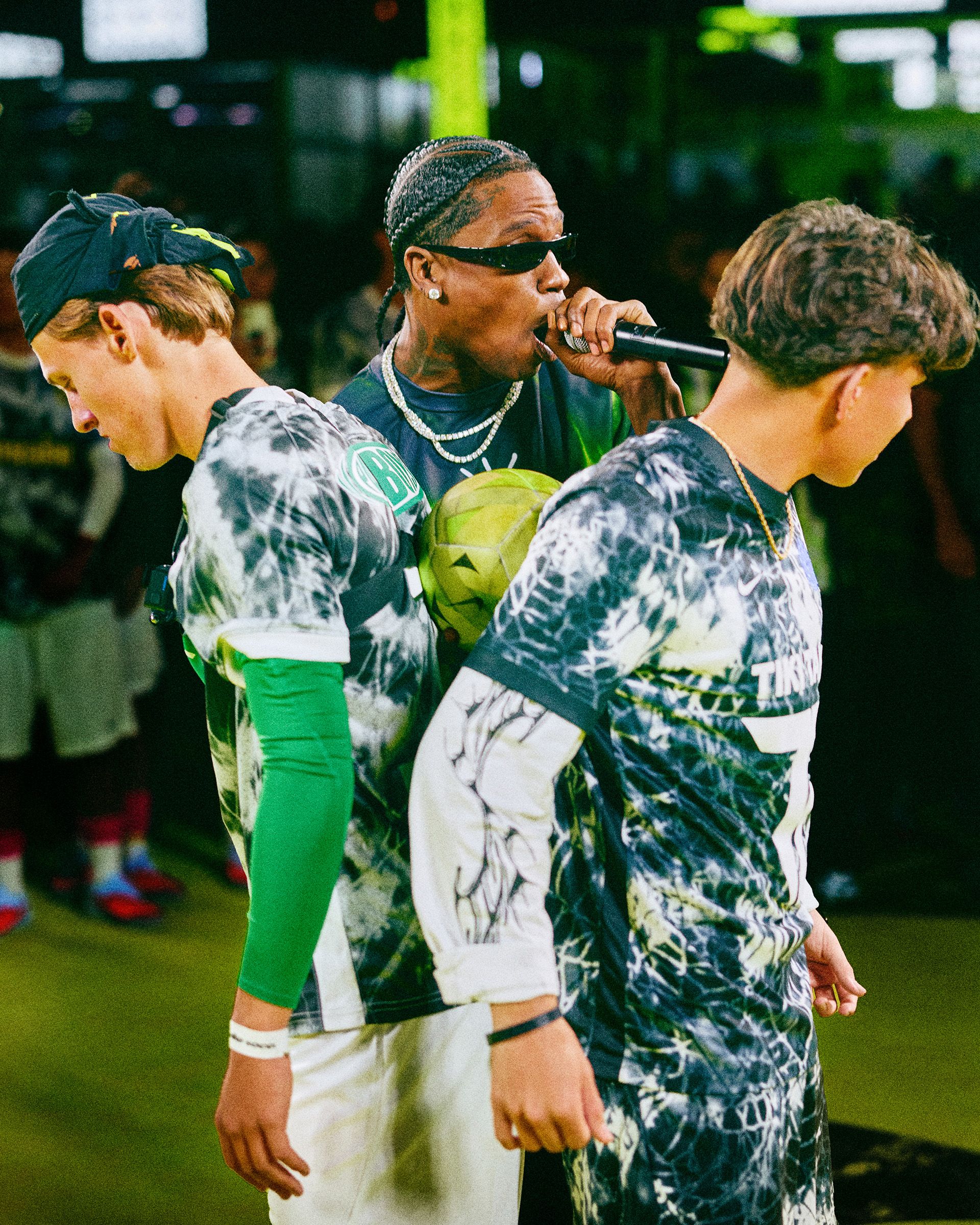 A performer with a microphone stands between two young athletes wearing sports uniforms during an indoor event.