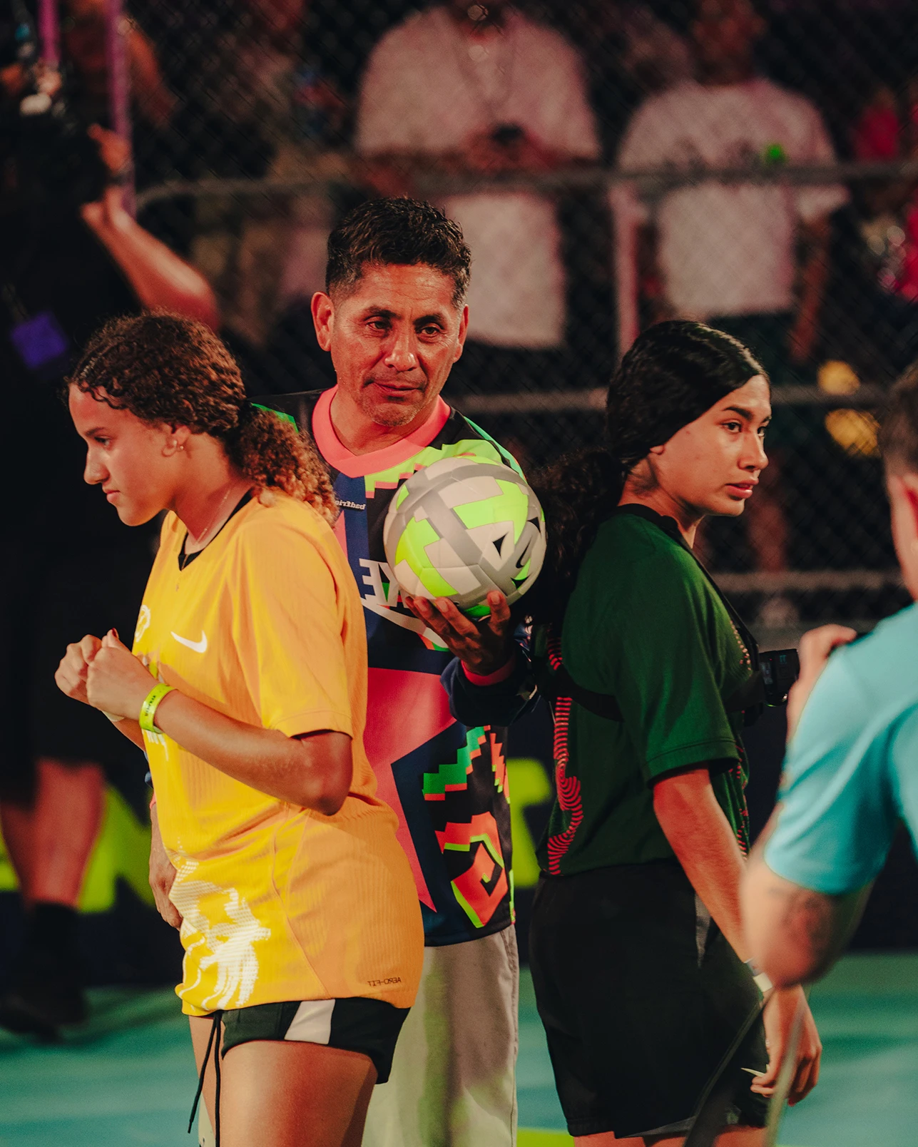 Three people stand on an indoor soccer court; one holds a soccer ball, while the other two in sports uniforms face away from each other. Spectators watch from behind a chain-link fence.