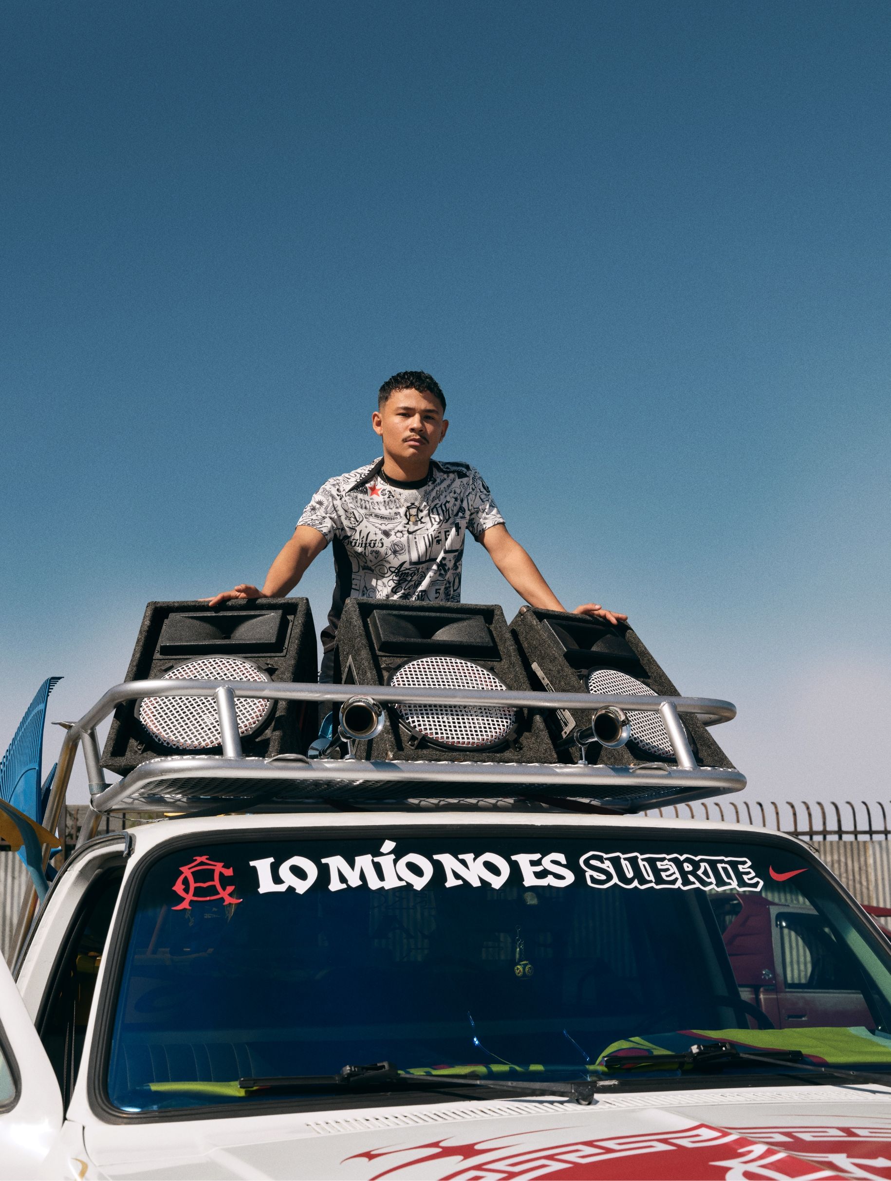 A young man stands on top of a vehicle roof beside large speakers. The windshield has a sign that reads "LO MÍO NO ES SUERTE." A clear blue sky is in the background.