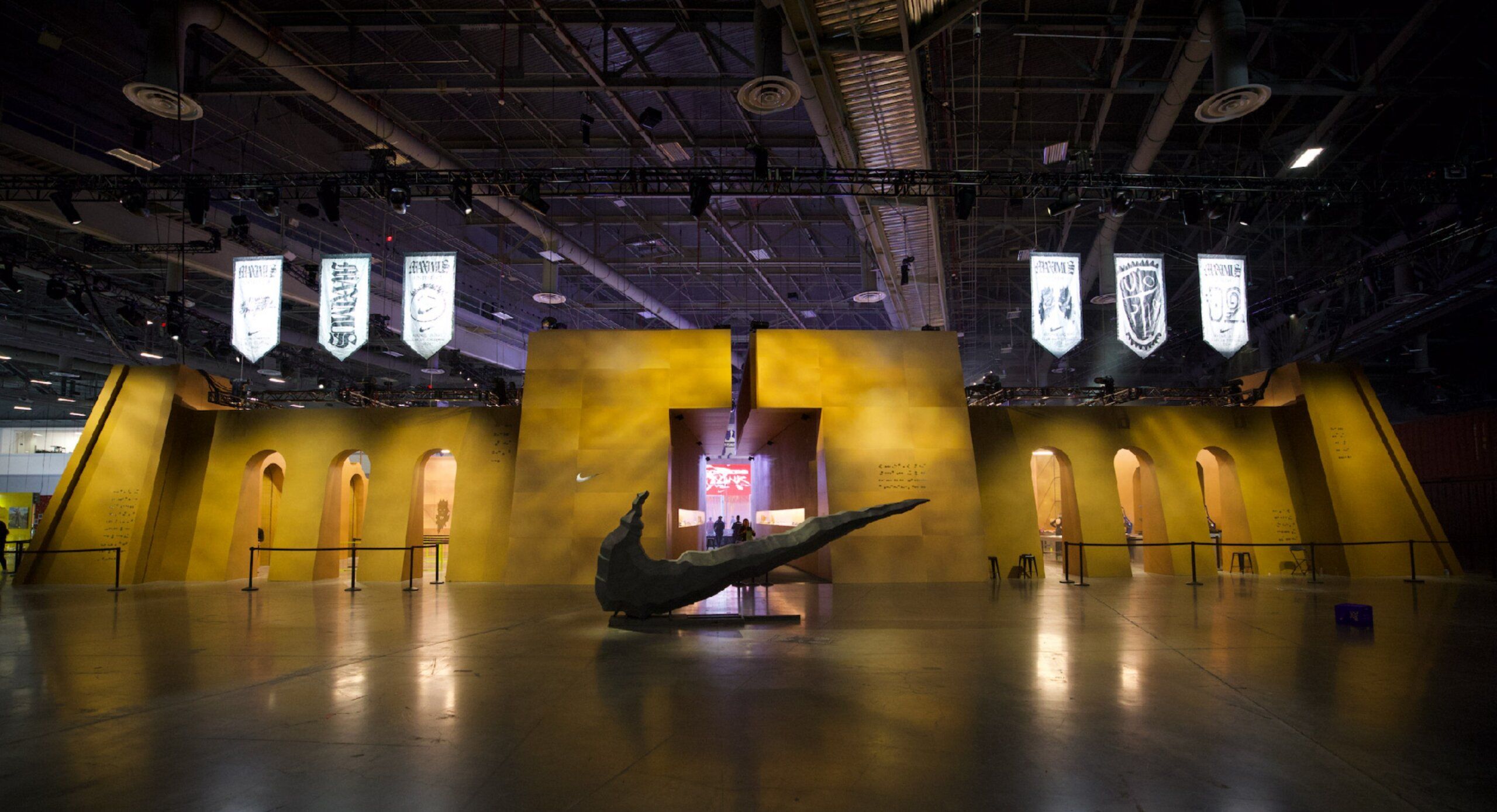 A large indoor installation with a dark curved sculpture in front of a yellow structure featuring arched doorways and hanging banners.