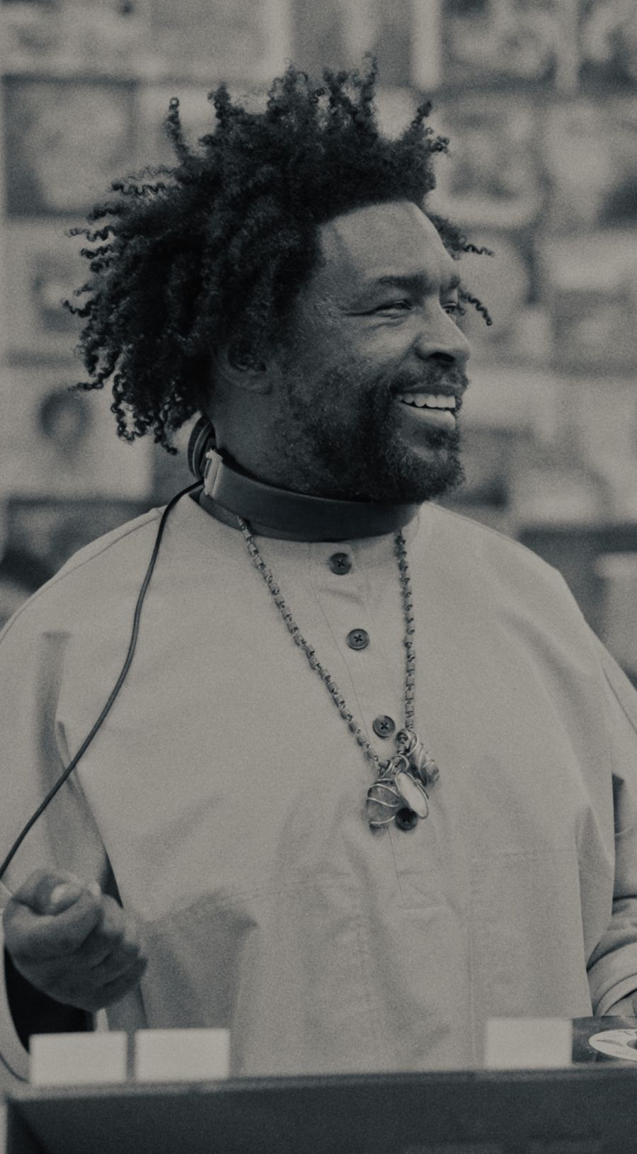 A man with curly hair, a beard, and a mustache smiles while wearing a light-colored shirt, beaded necklaces, and headphones around his neck.
