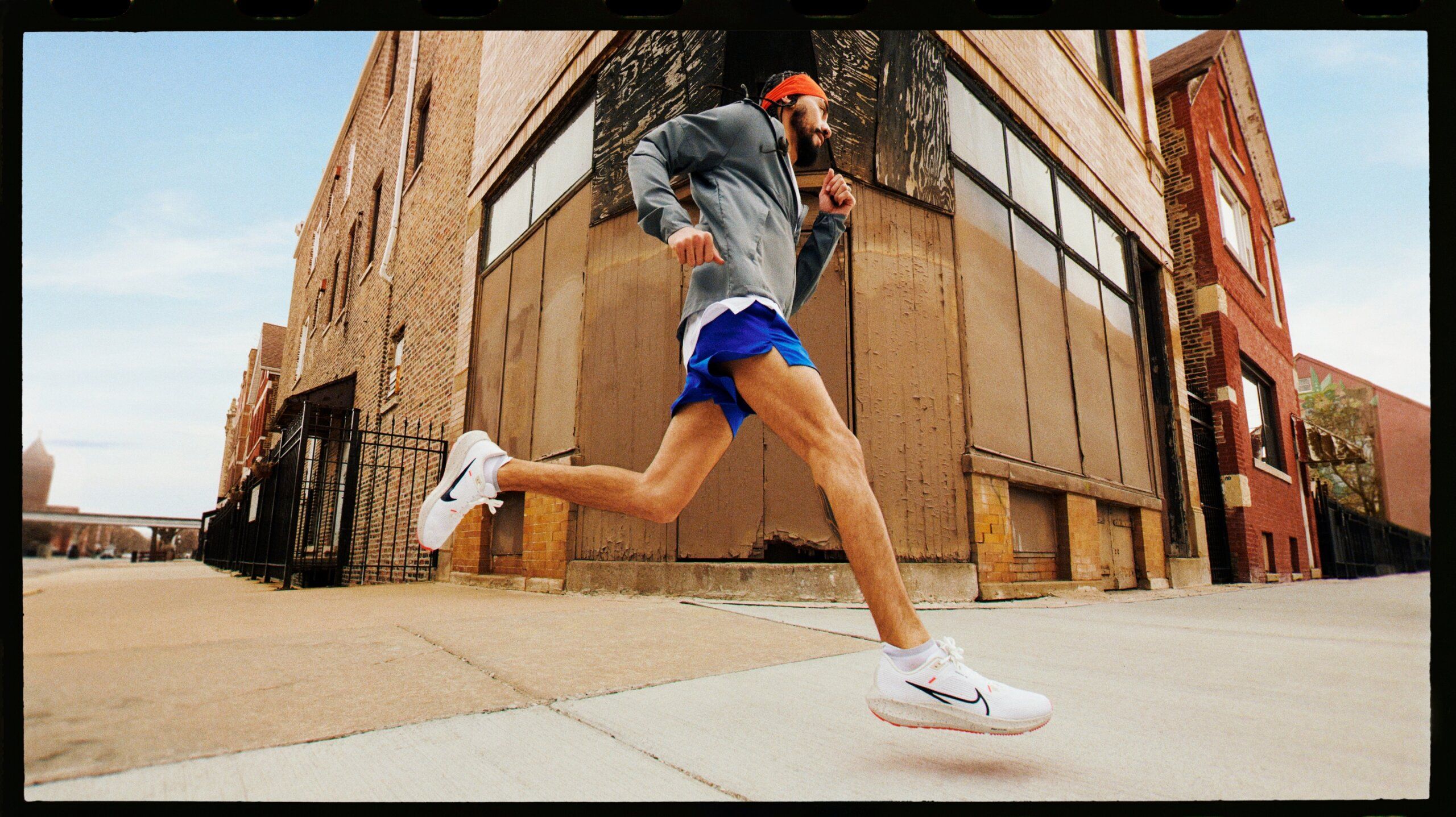 Main running full stride on city sidewalk wearing orange Nike bandana, gray warm up jacket, blue running shorts and white Nike Pegasus running shoes.