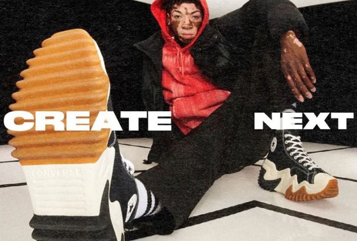 Person in a red hoodie and black jacket sits on the floor, one leg extended forward showing a sneaker with a thick, ridged sole. Text reads "CREATE NEXT.