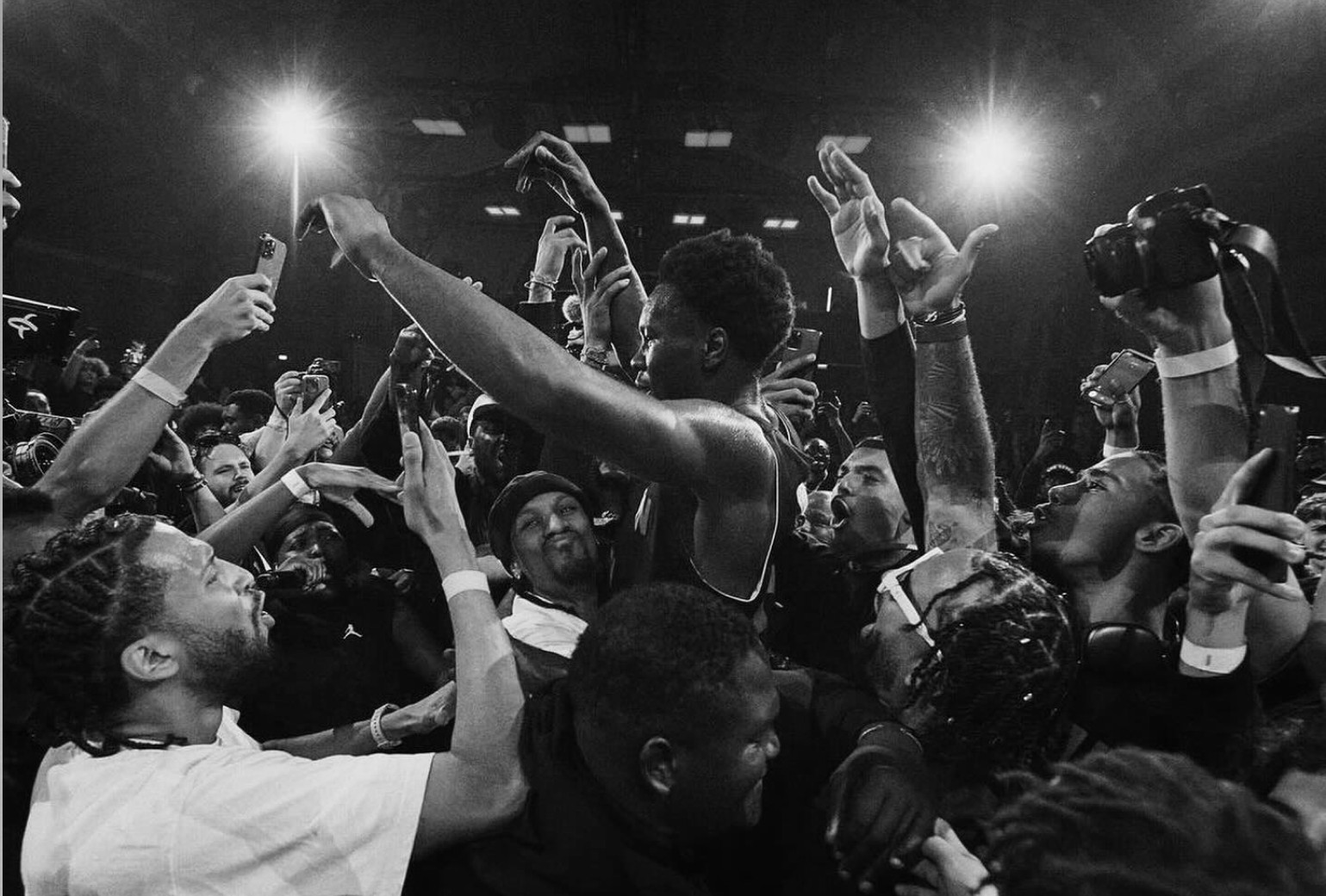 A basketball player is lifted and surrounded by an excited crowd of fans, with many reaching out and taking photos in a dimly lit indoor setting.