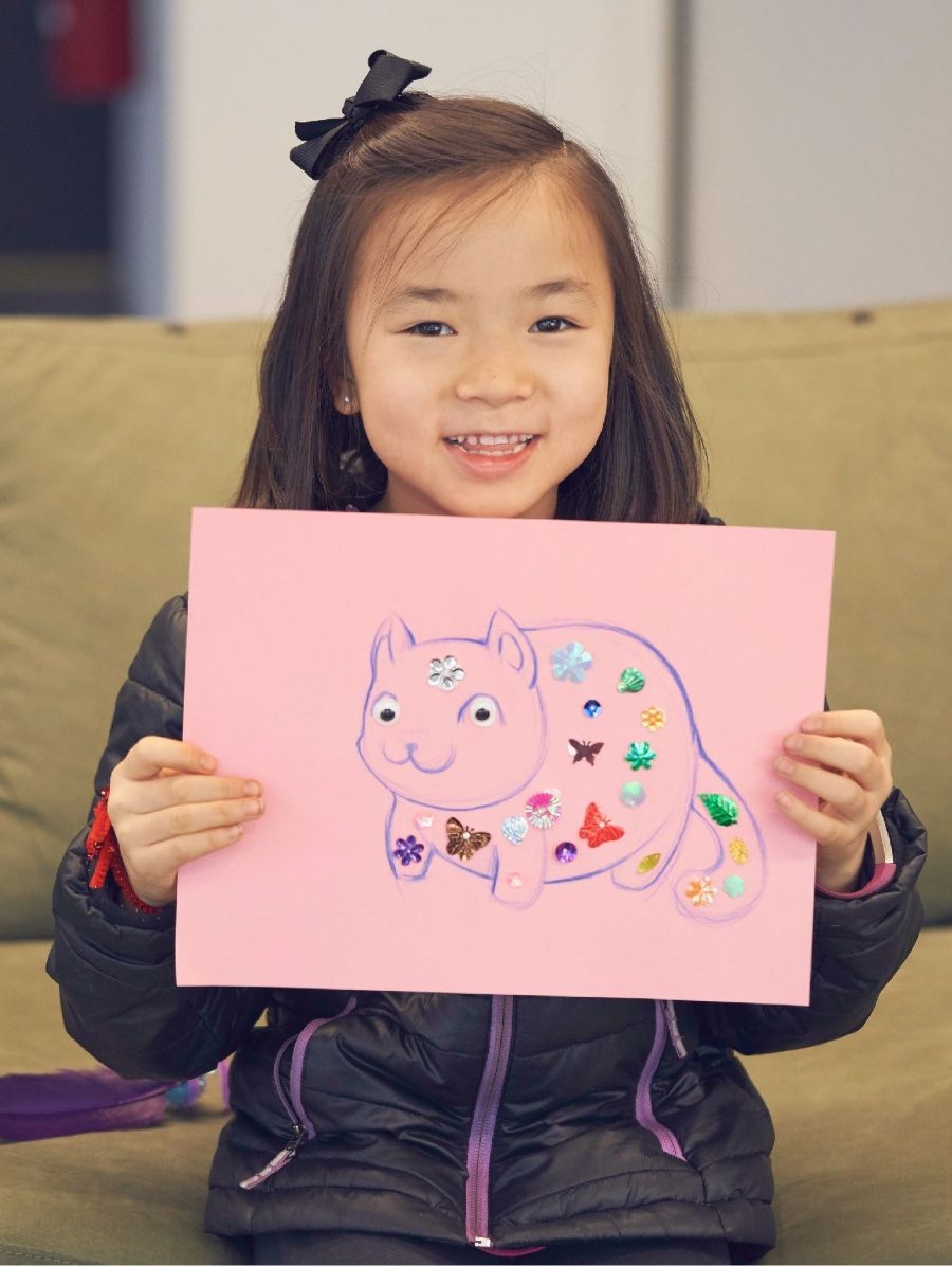 Young girl smiling holding up drawing of cat with stickers covering drawing.