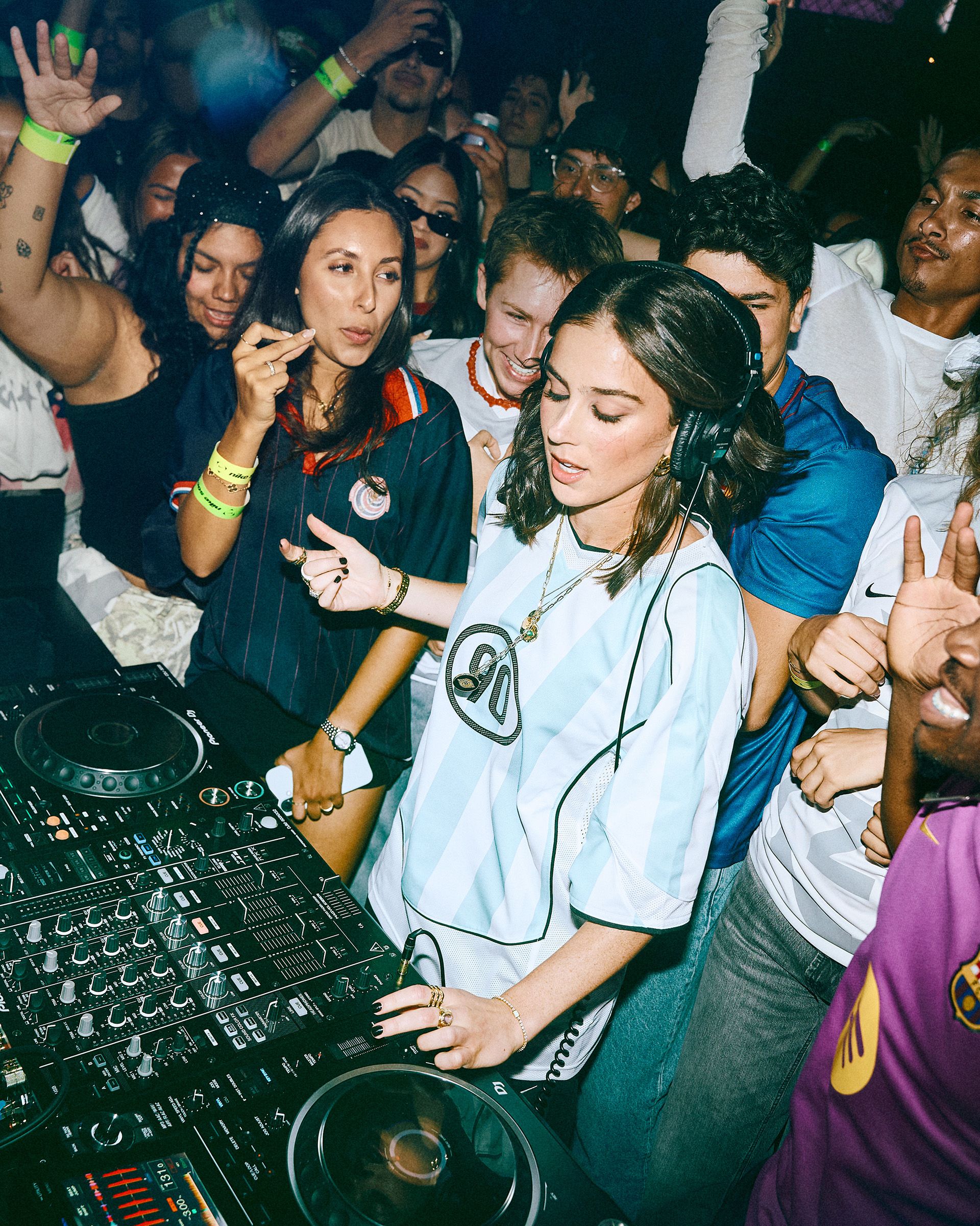 A DJ wearing headphones performs at a crowded party, surrounded by people dancing and enjoying the music.