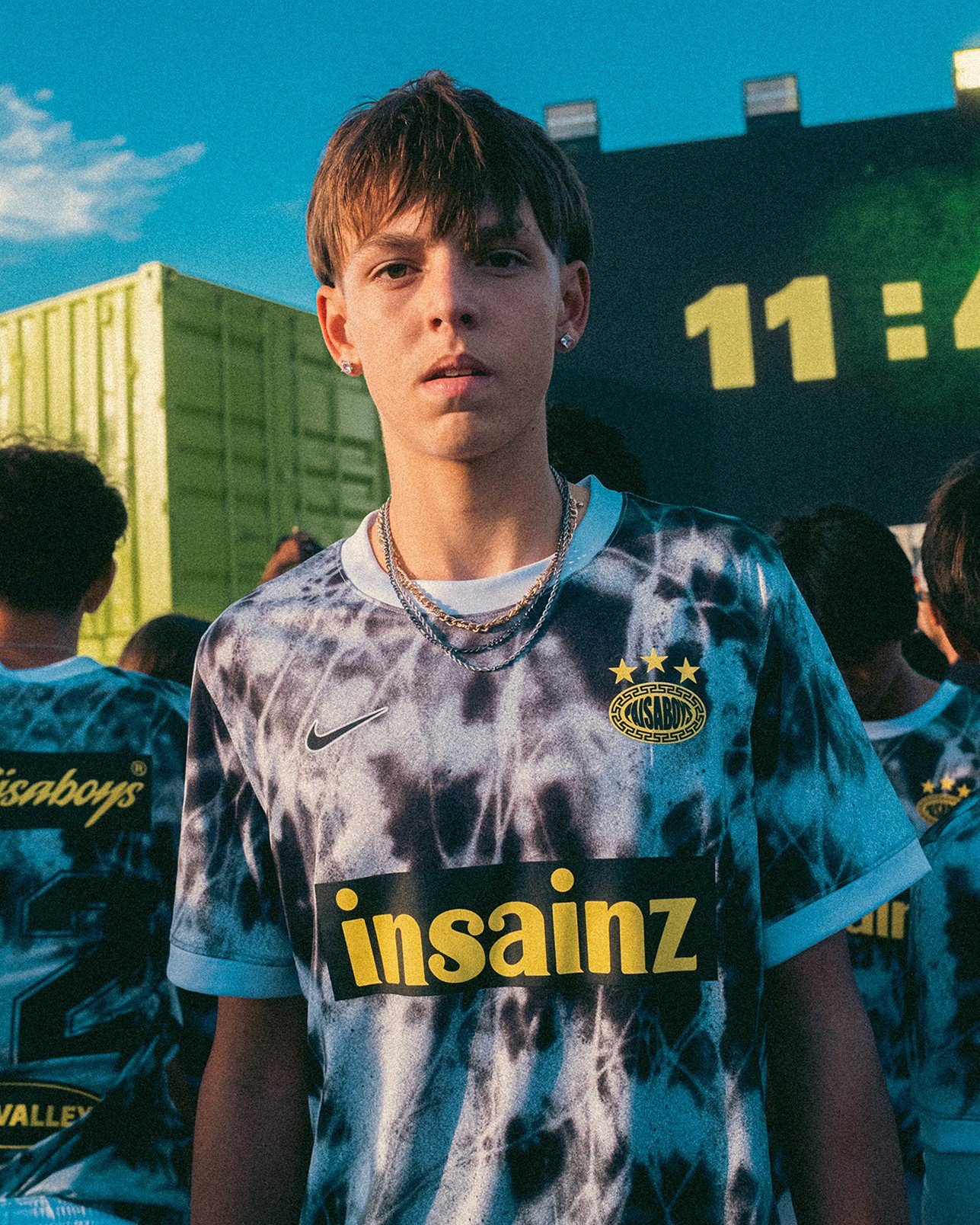 Teen boy wearing a blue and black tie-dye soccer jersey stands outdoors among a group, with containers and a digital clock in the background.