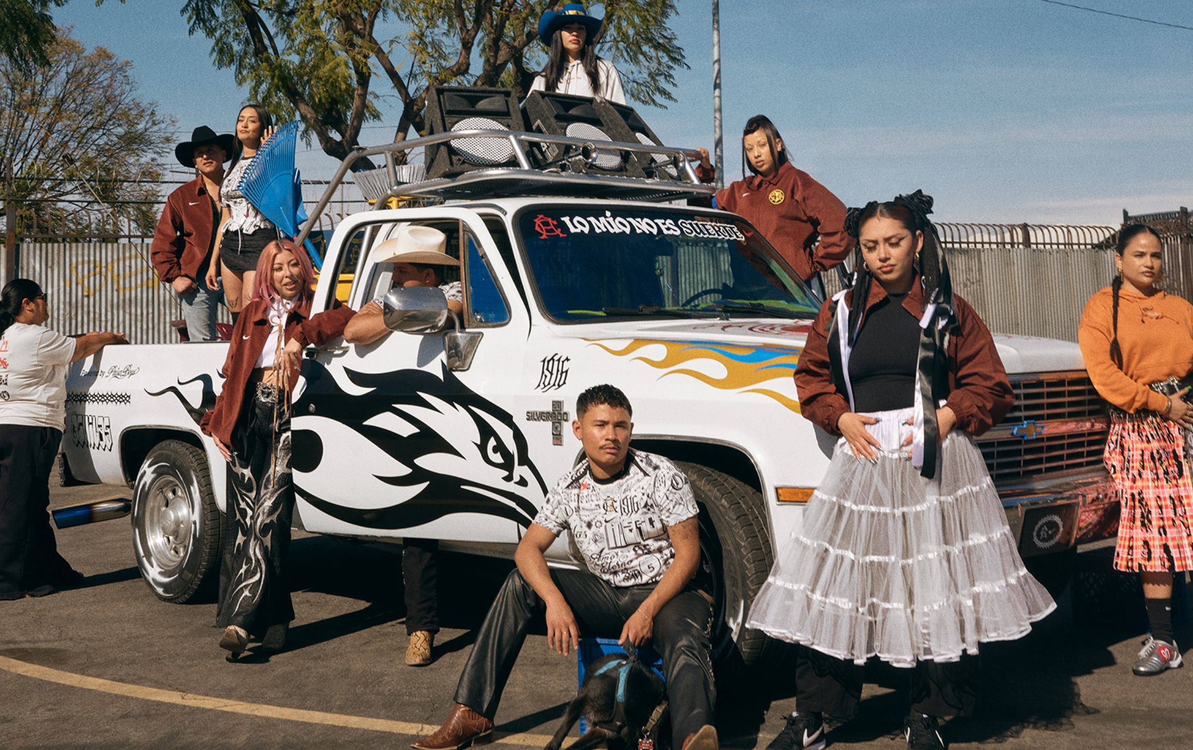 A group of people poses around a white pickup truck with black and blue flame decals, parked in an outdoor lot near a fence and trees.