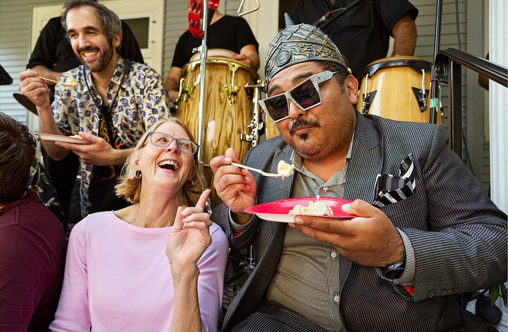 A woman and a man in a suit eat cake and smile at an outdoor gathering, with musicians and drums in the background.