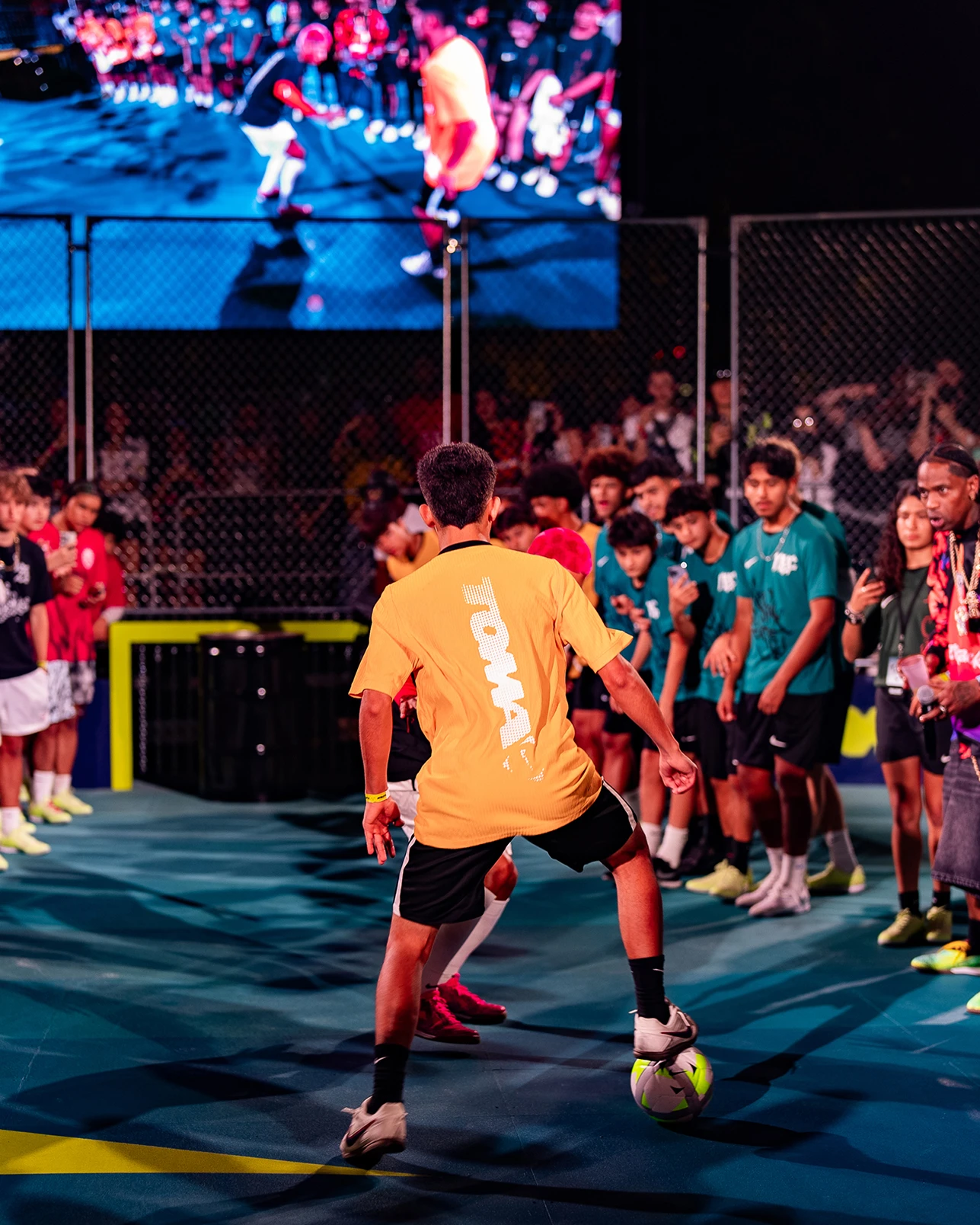 A soccer player in a yellow shirt dribbles a ball on an indoor court, surrounded by spectators and other players, with a large screen displaying the game behind them.