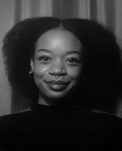 A person with natural curly hair, a nose ring, and hoop earrings smiles slightly while facing the camera in a black and white photo.