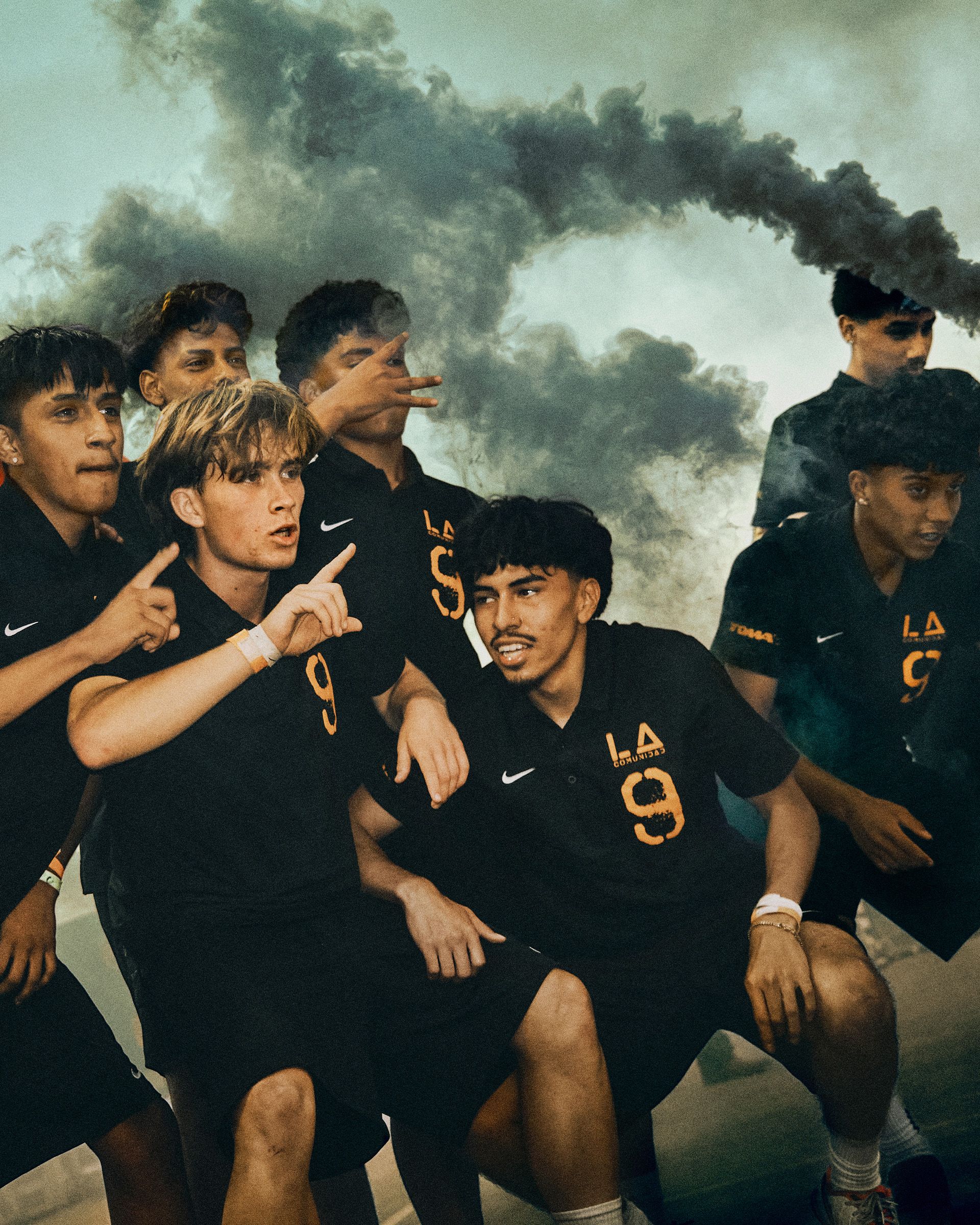 A group of young men in matching black sports uniforms pose together outdoors, with smoke clouds rising in the background.