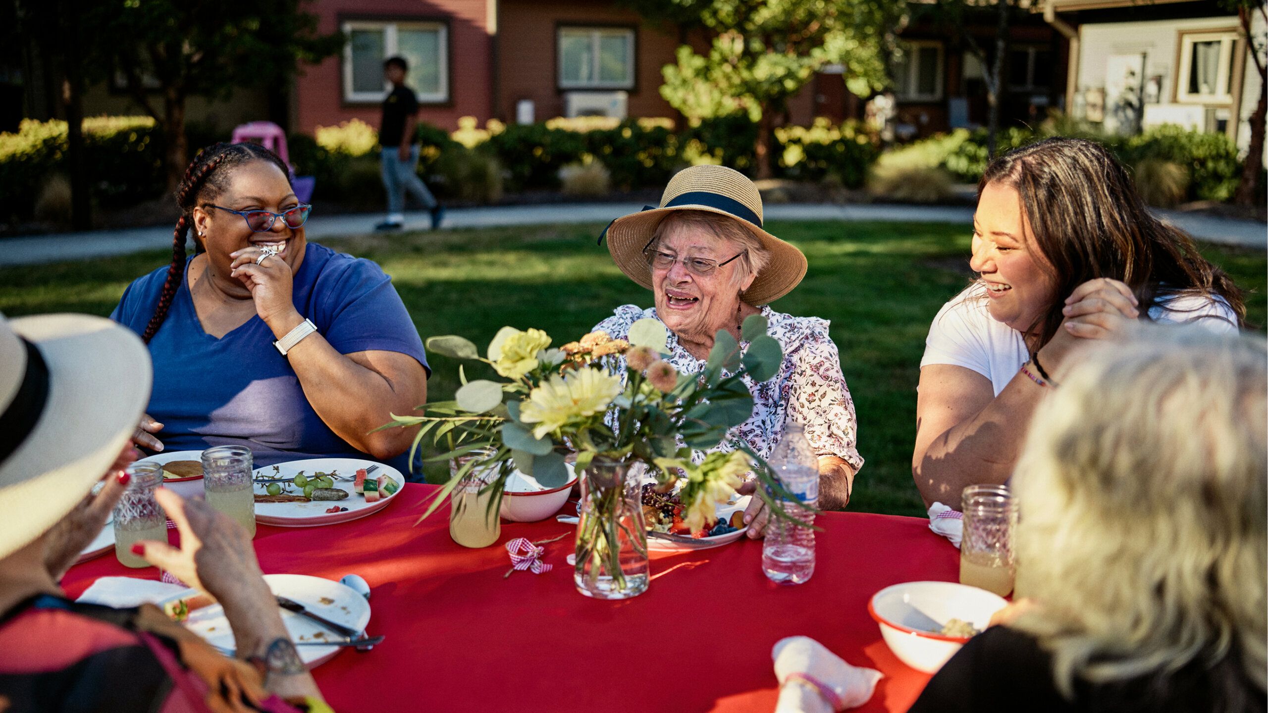 A group of people sit outdoors around a red table, sharing a meal, talking, and laughing on a sunny day.