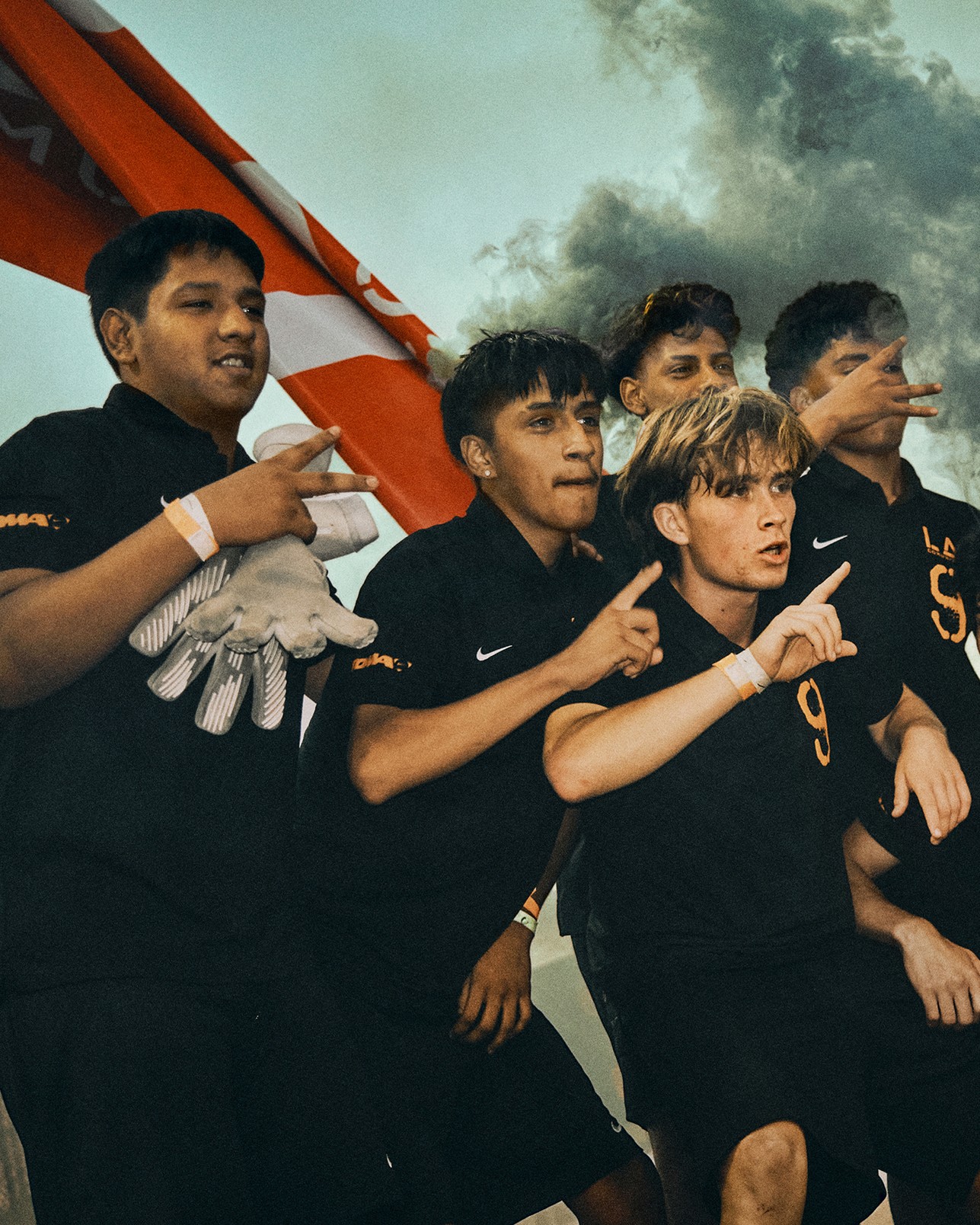 Five young men in black uniforms pose together outdoors, holding up one finger each, with a red and white flag and cloudy sky in the background.