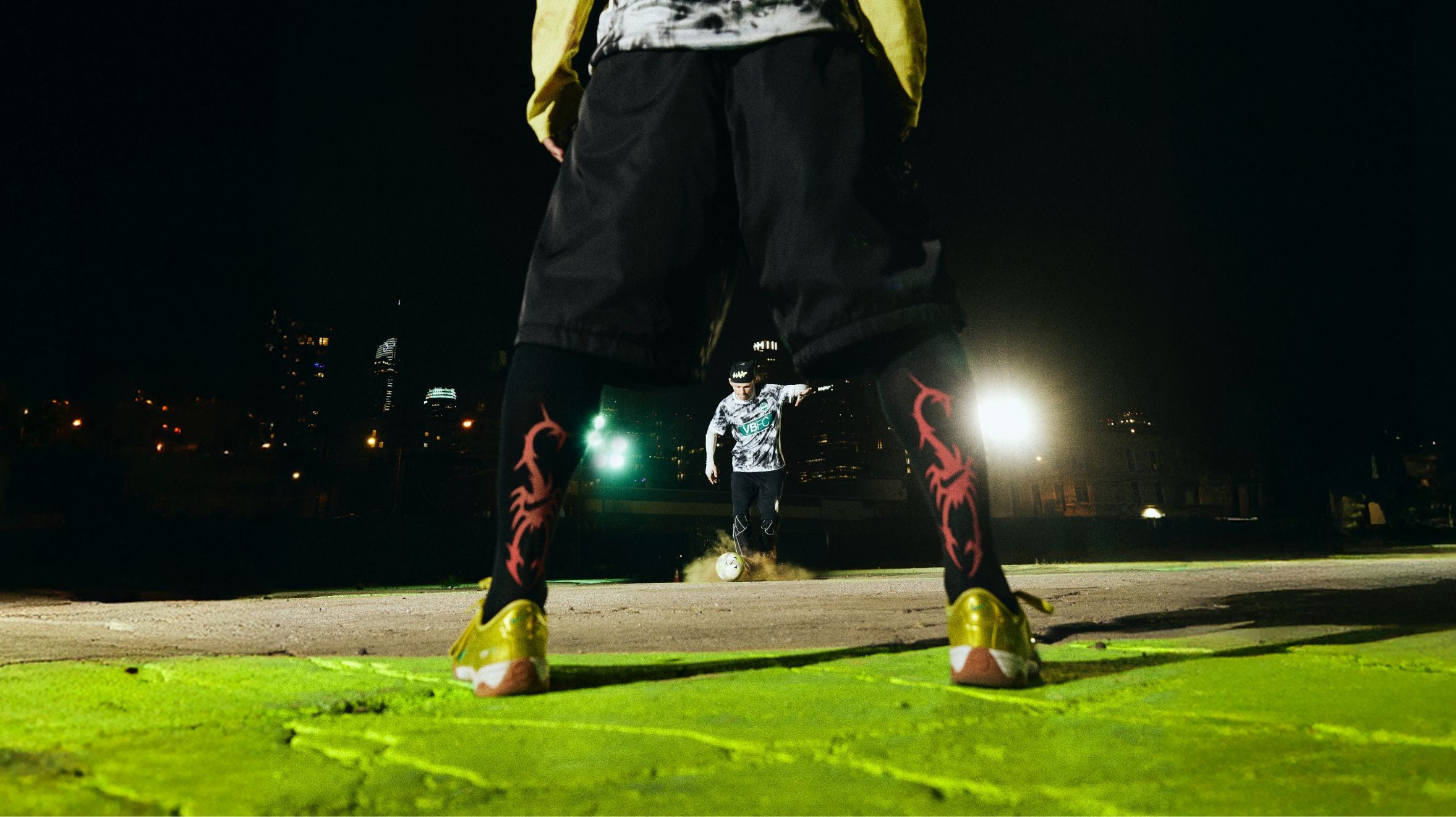 A soccer player prepares to take a penalty shot at night, viewed from behind the goalkeeper standing with legs apart on a green-lit surface.