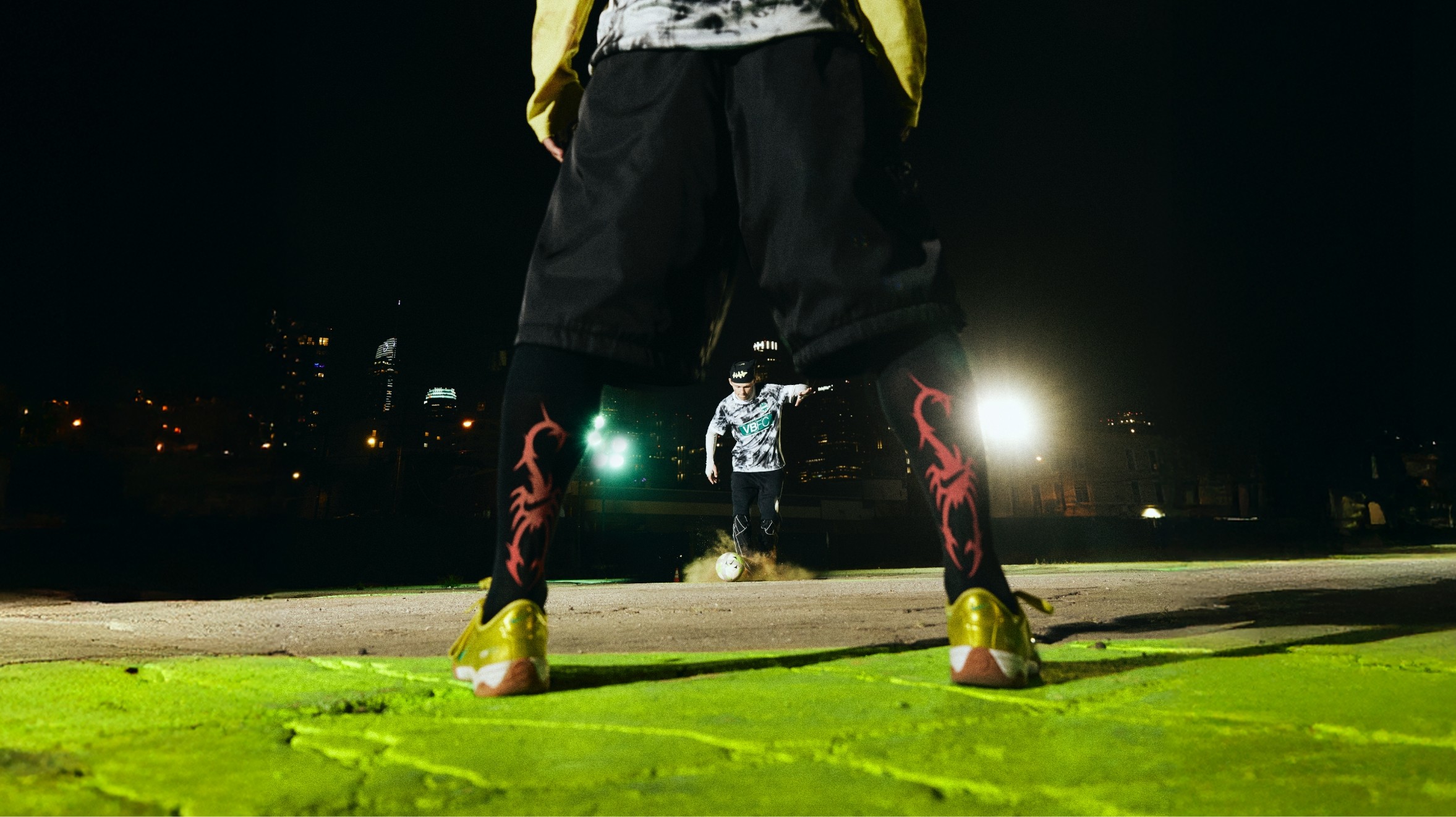 A soccer player prepares to take a penalty shot at night, viewed from behind the goalkeeper standing with legs apart on a green-lit surface.