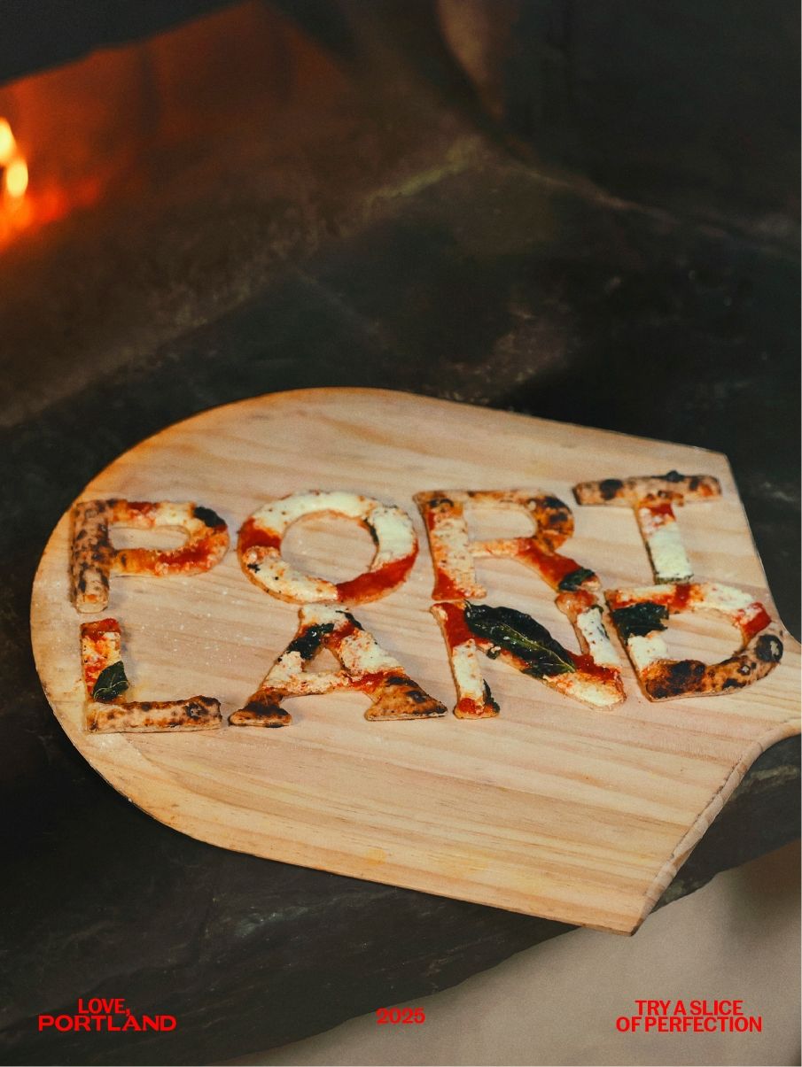 A wooden pizza peel with pizza cut out to spell "PORTLAND," placed in front of a pizza oven. Text at the bottom reads "LOVE PORTLAND 2020" and "TRY A SLICE OF PERFECTION.