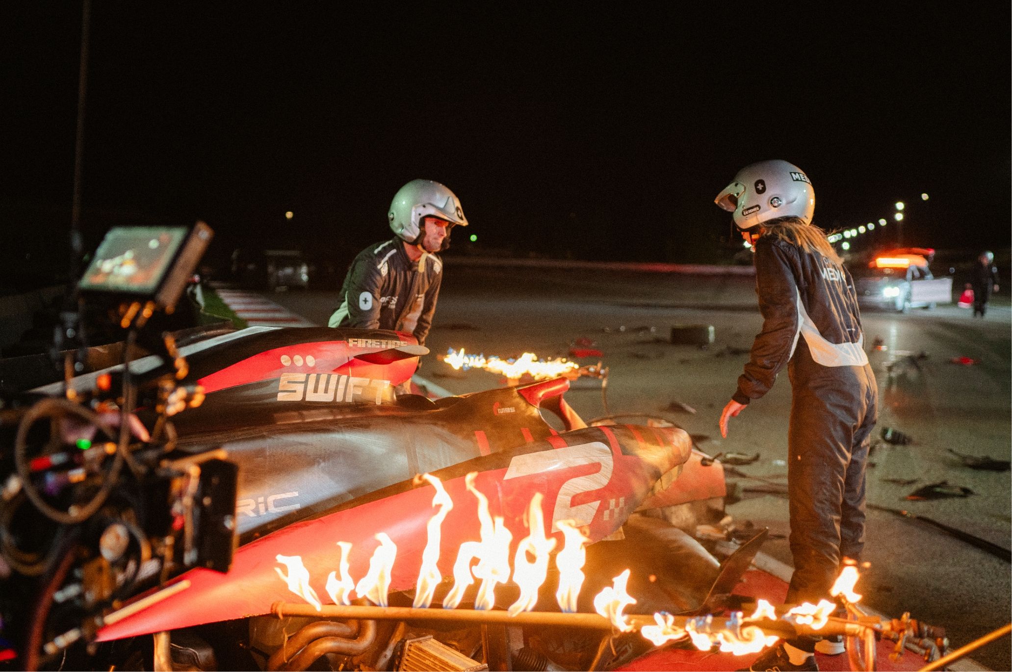 Two people in racing suits stand near a burning race car at night, with film equipment visible on the left.