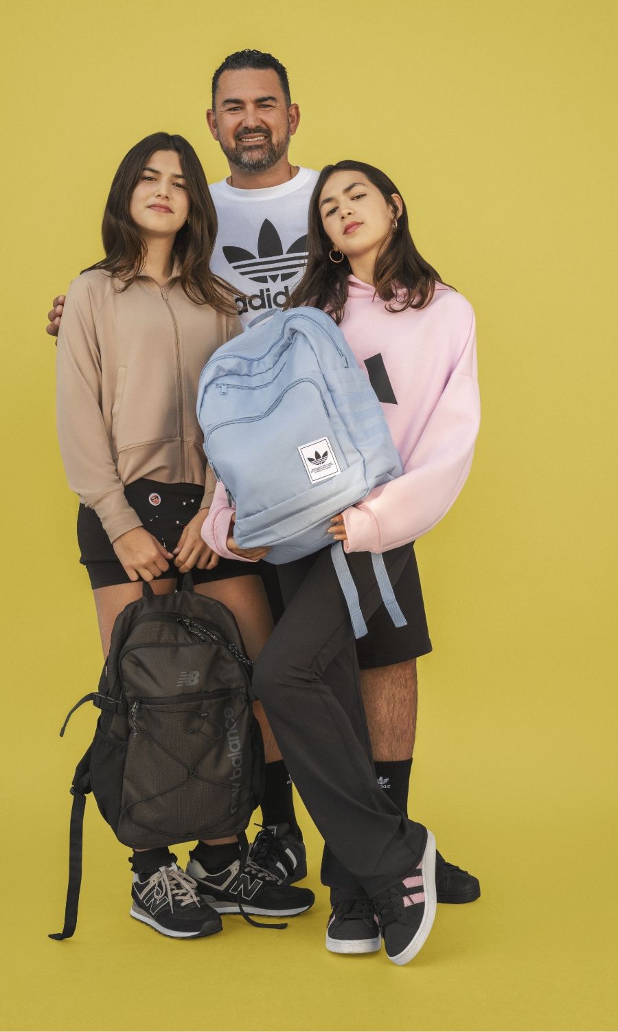 A man stands behind two teenage girls holding backpacks, all wearing casual sportswear, against a plain yellow background.