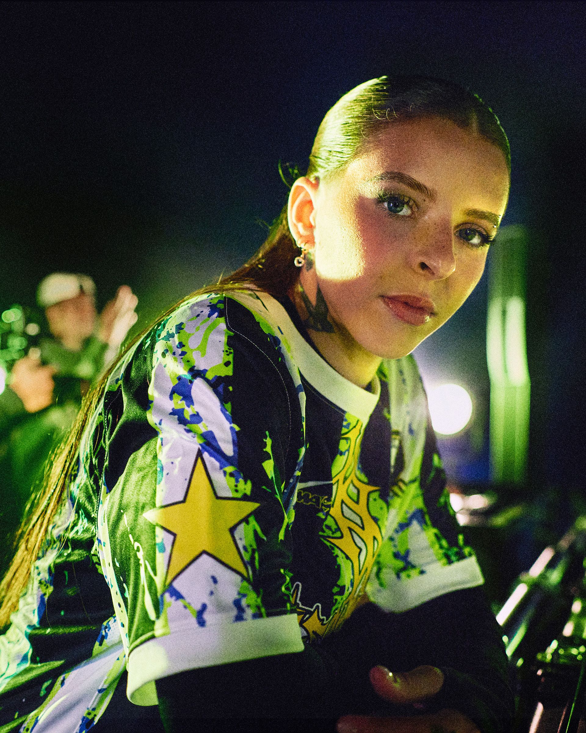 A person with long dark hair and a colorful shirt with yellow stars looks at the camera in a dimly lit setting; blurred figures and lights appear in the background.