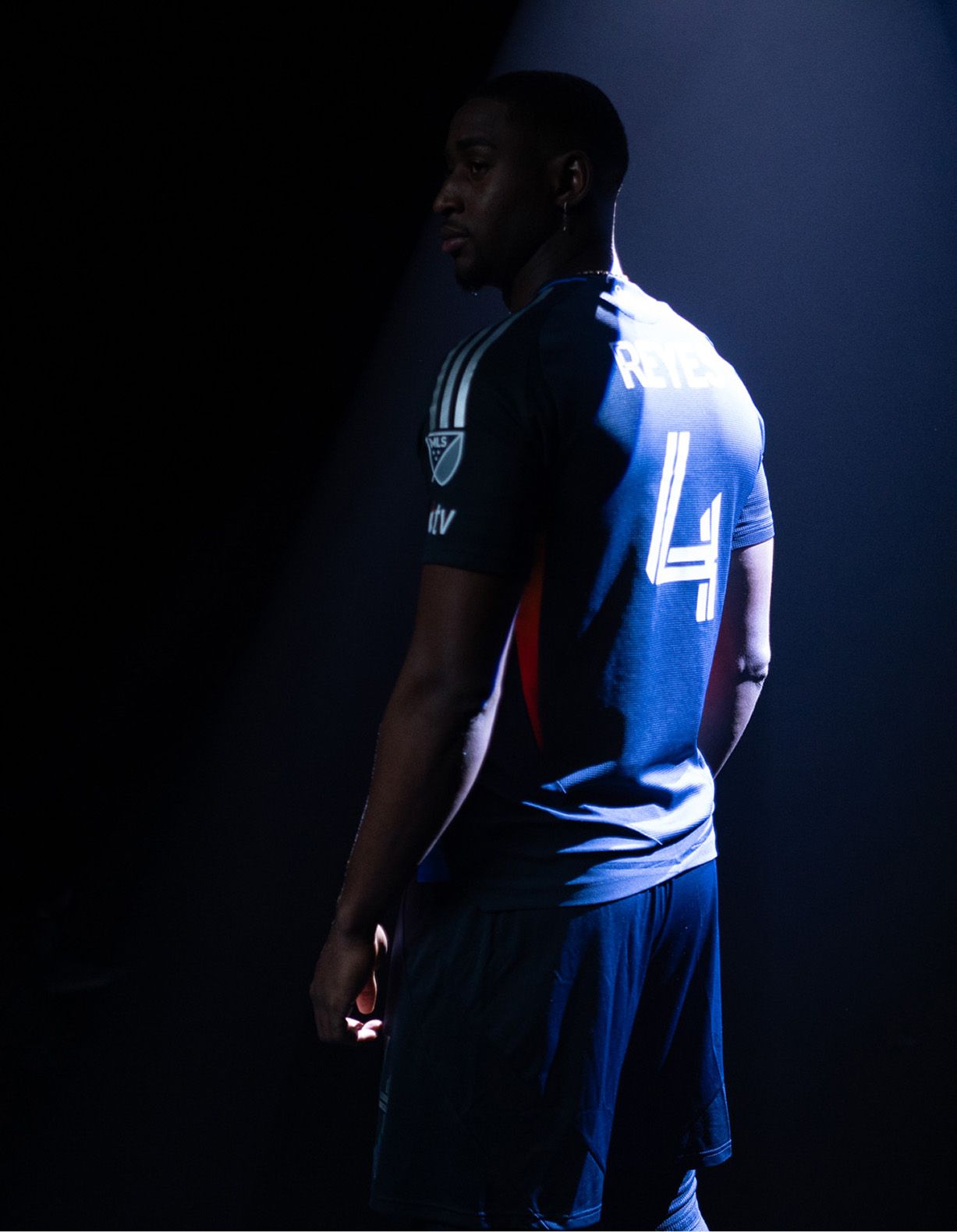 A soccer player wearing a dark jersey with the name "REYES" and number 4 stands under a spotlight, facing slightly away from the camera.