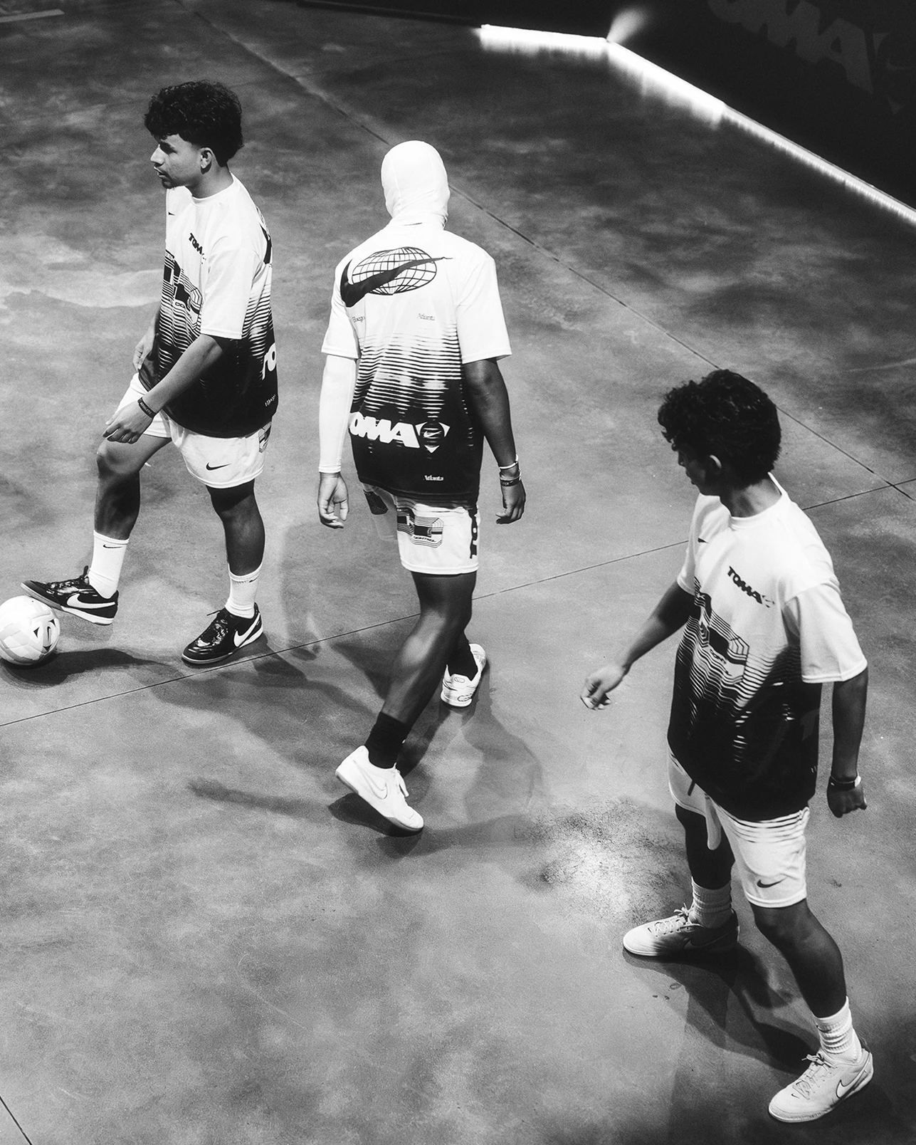 Three people in sports uniforms stand on an indoor court with a soccer ball nearby; one person has a shirt covering their head.