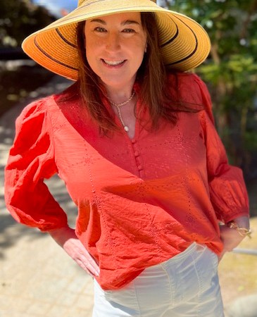 Woman wearing a wide-brimmed straw hat, red blouse, and white shorts stands outdoors, smiling at the camera with one hand on her hip.