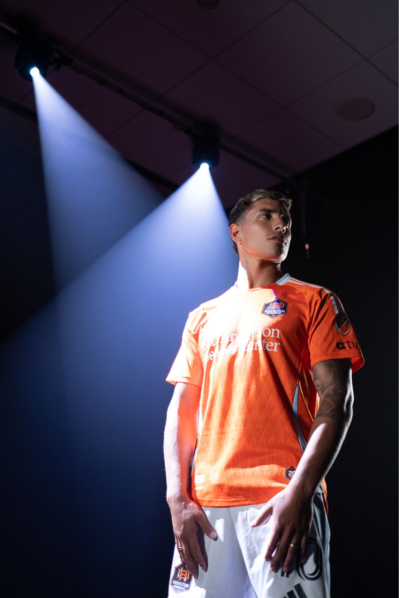 A soccer player wearing an orange jersey and white shorts stands under spotlights, looking to the side in a dimly lit indoor setting.