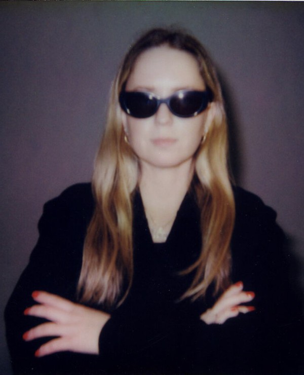 A person with long blond hair wearing dark sunglasses and a black outfit stands with arms crossed against a plain background.