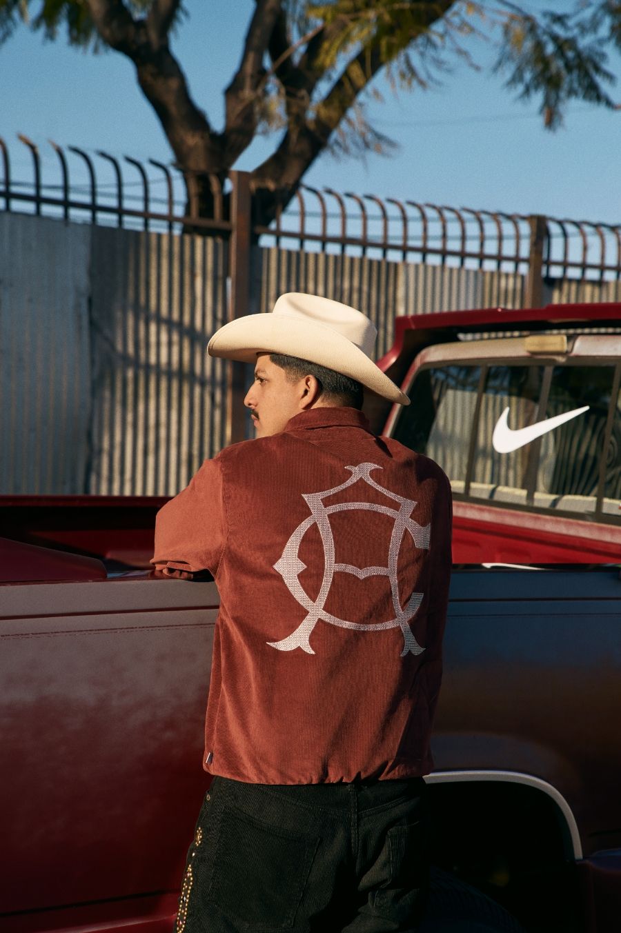 A man in a cowboy hat and brown shirt with a large white symbol on the back leans against a red truck, with a fence and tree in the background.