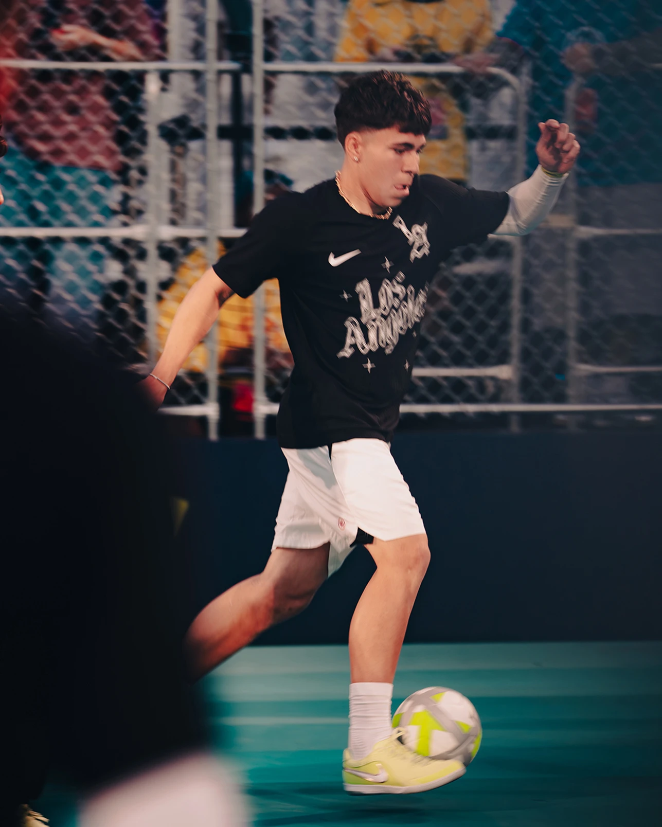 A young man in a black shirt and white shorts dribbles a soccer ball on an indoor court. Spectators are visible behind a chain-link fence.