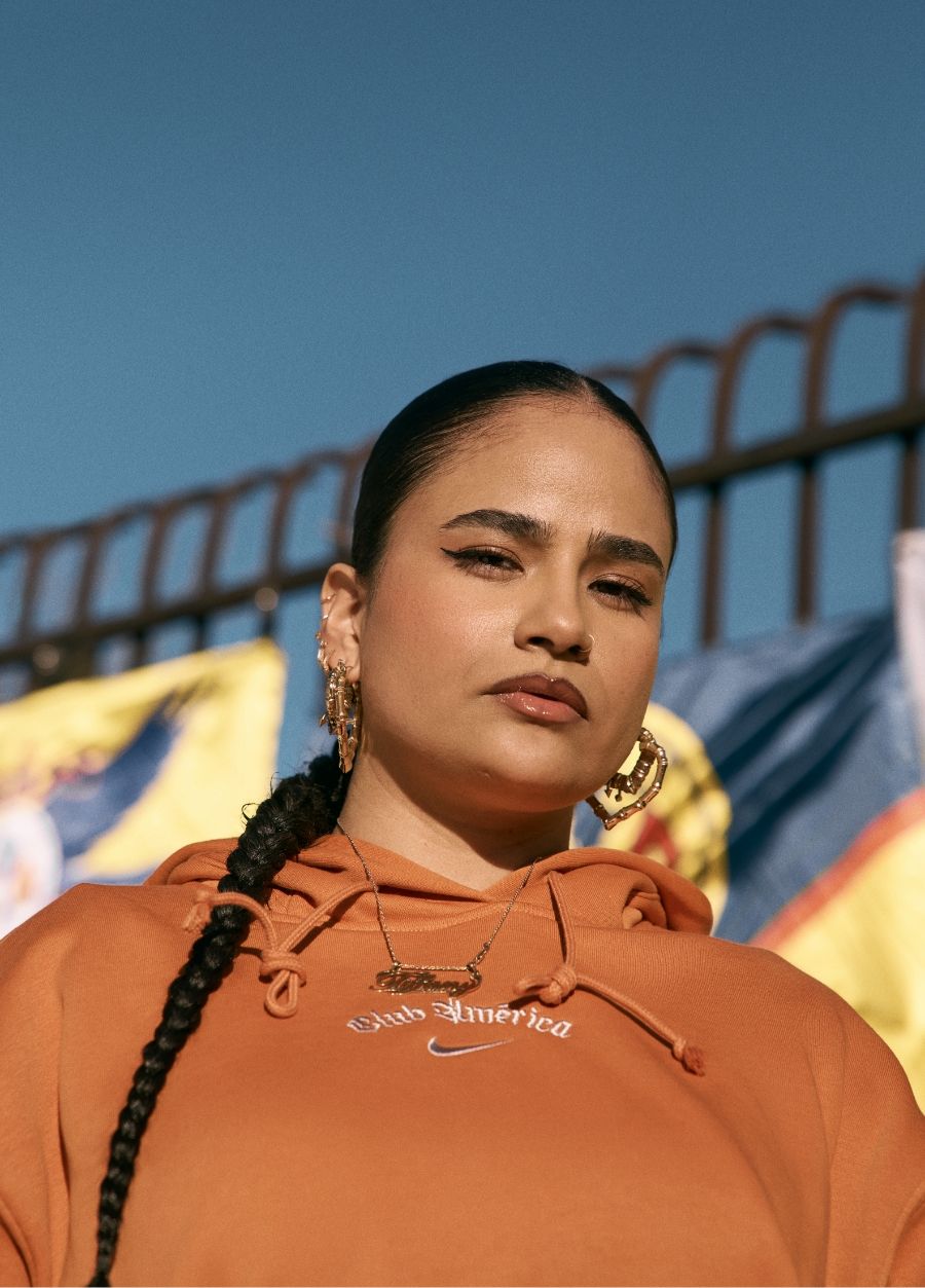 A woman with a long braid and hoop earrings wears an orange hoodie and gold necklaces, standing outdoors in front of colorful flags and a metal fence.